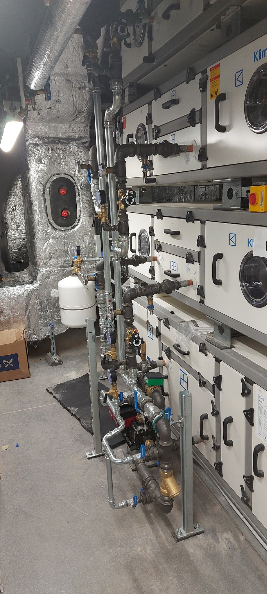 Rozbudowana instalacja z rur i zaworów przy systemie wentylacyjnym Klivent EKO, widoczne manometry i pompa obiegowa, rury z izolacją termiczną.