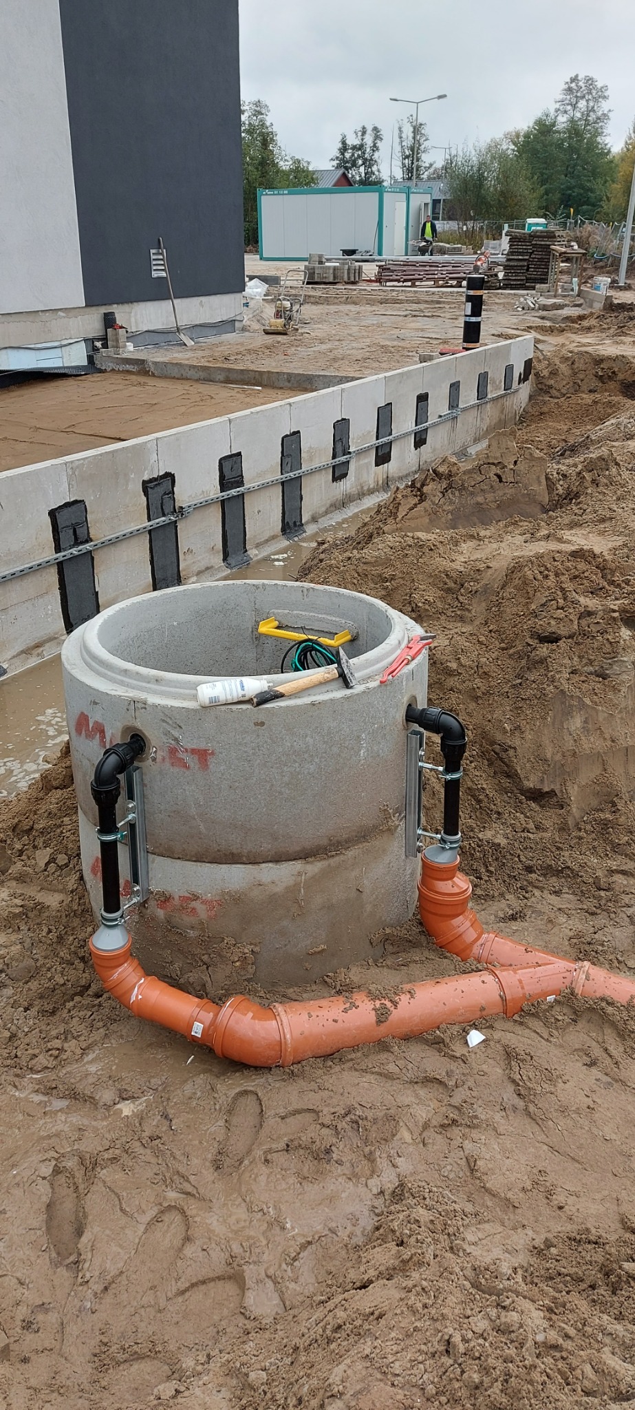 Betonowy krąg studzienny z podłączonymi rurami kanalizacyjnymi z tworzywa sztucznego, leżące narzędzia na górze, w tle fundamenty budynku i teren budowy.