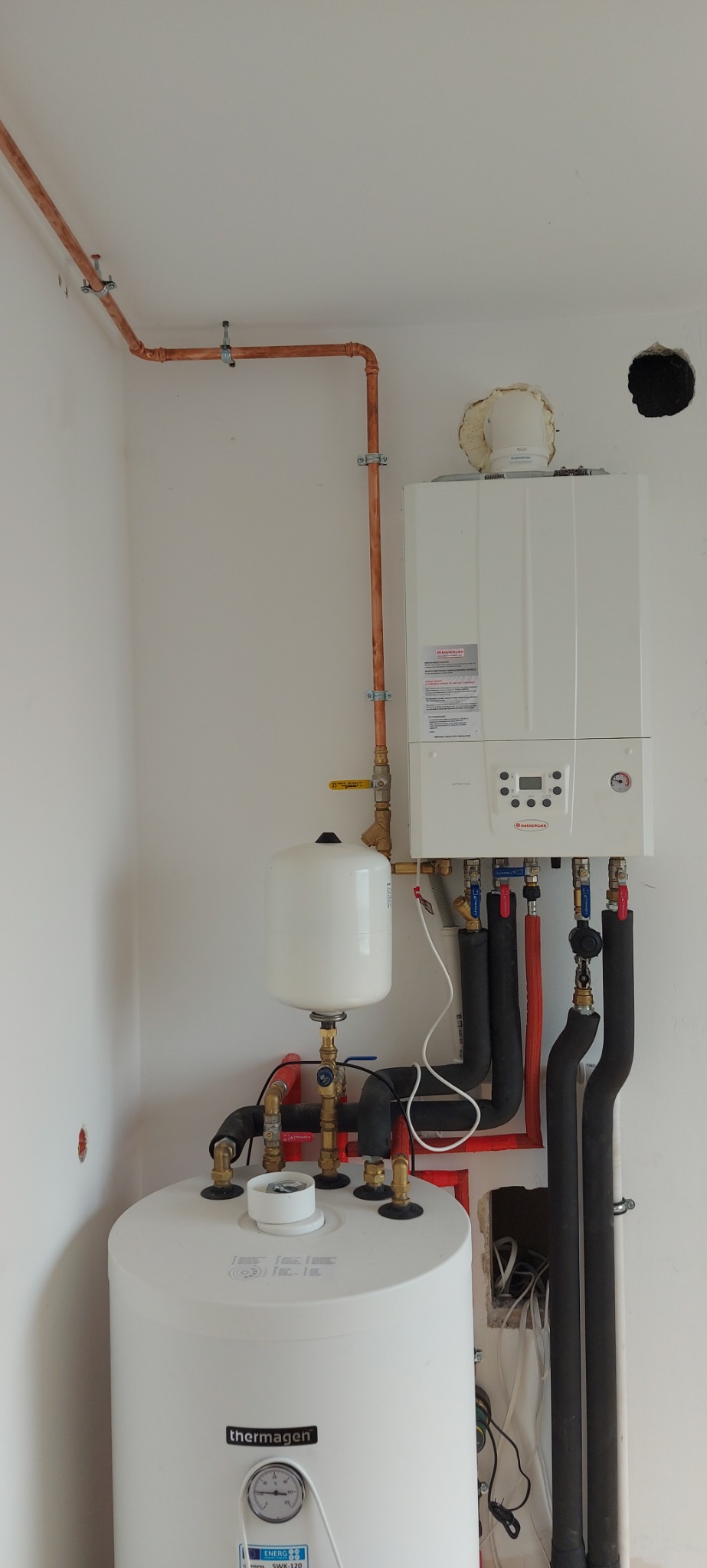 Nowoczesny, biały piec gazowy dwufunkcyjny marki Immergas z podłączonymi rurami miedzianymi i czarnymi rurami izolowanymi, zbiornik wyrównawczy i zasobnik ciepłej wody Thermagen, zamontowany...