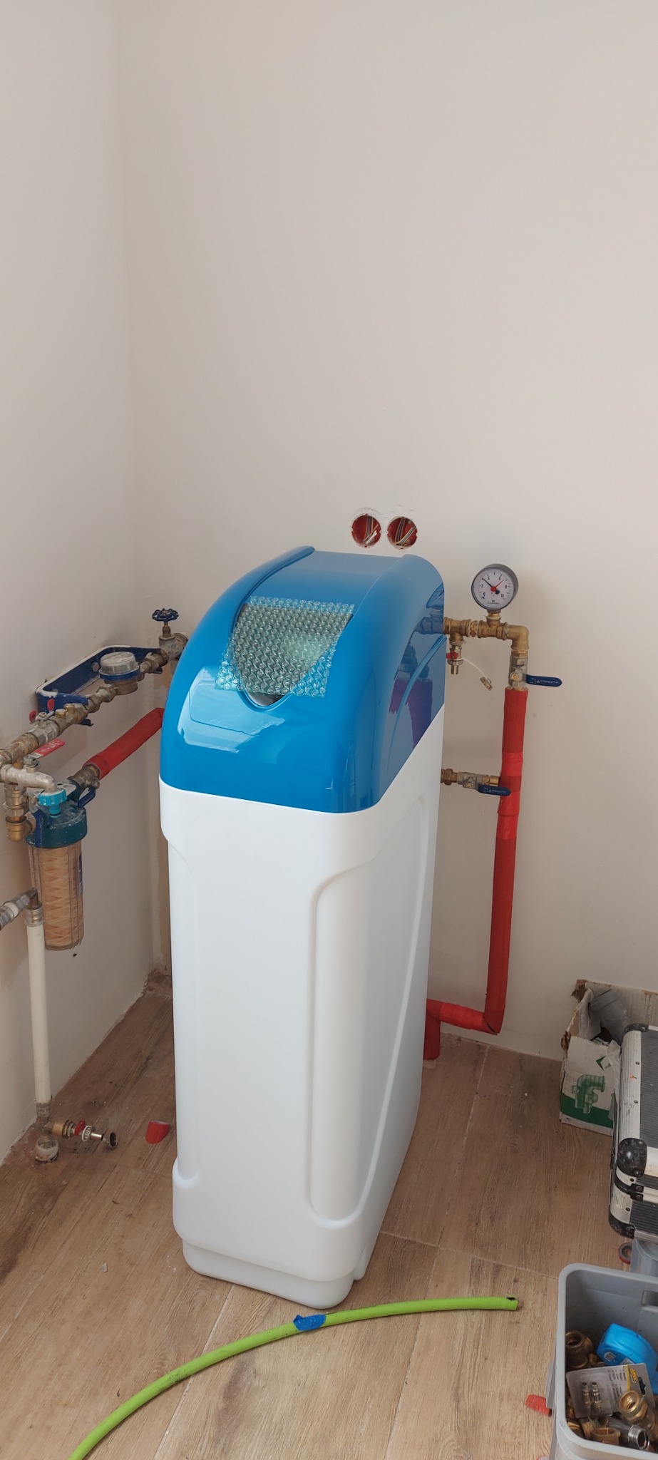 Nowa stacja uzdatniania wody z niebieską pokrywą, podłączona do instalacji hydraulicznej w pomieszczeniu z panelami podłogowymi.