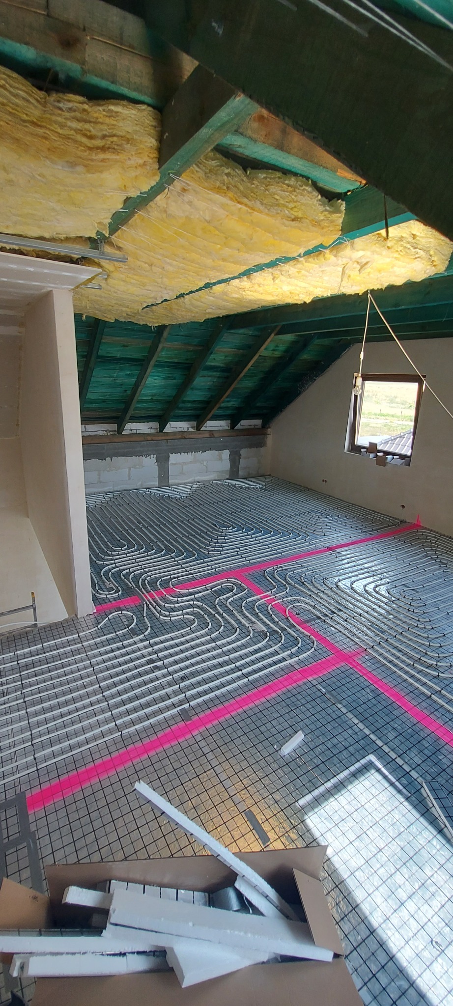 Montaż ogrzewania podłogowego w nowym budynku: białe rury ułożone na siatce, zaznaczone różową farbą, widoczna izolacja stropu i fragment okna.