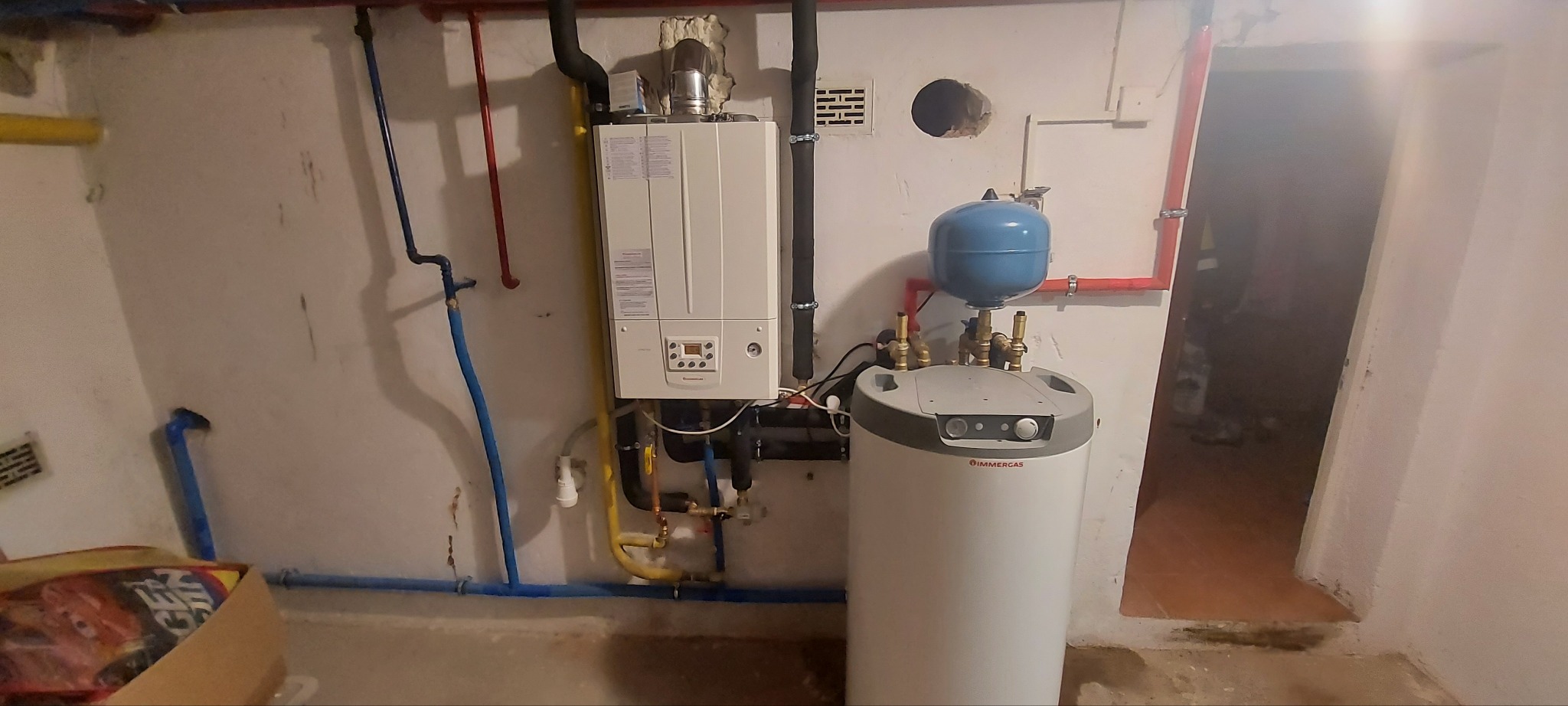Kocioł gazowy Immergas z podłączonym zasobnikiem ciepłej wody, widoczne rury w kolorach żółtym, niebieskim i czerwonym, w tle ściana z niedoskonałościami.