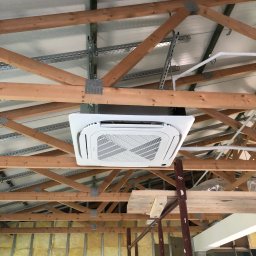 Ecowent - Biała kaseta klimatyzacyjna zamontowana pod drewnianą konstrukcją dachu, widoczne metalowe profile mocujące i ocieplenie ścian wełną mineralną.