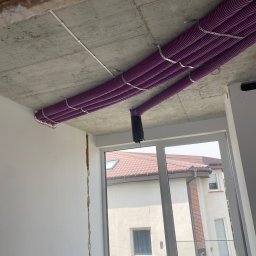 Ecowent - Fioletowe rury rekuperacji zamontowane pod surowym sufitem w budynku w Jesionce, widoczne przez okno sąsiednie budynki.