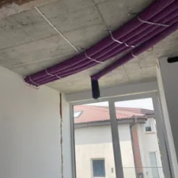 Fioletowe rury rekuperacji zamontowane pod surowym sufitem w budynku w Jesionce, widoczne przez okno sąsiednie budynki.