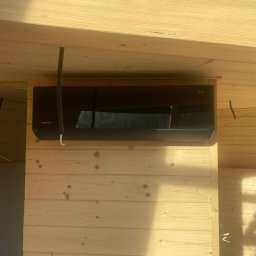 Ecowent - Czarna klimatyzacja Rotenso Teta zamontowana na drewnianej ścianie z boazerii, widoczne rury instalacyjne i surowe drewniane belki stropowe.