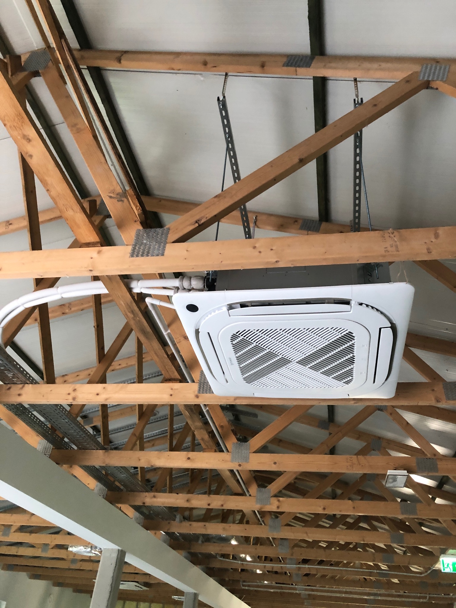 Biała, kasetonowa jednostka klimatyzacji zamontowana pod drewnianą konstrukcją dachu z widocznymi przewodami chłodniczymi w izolacji.