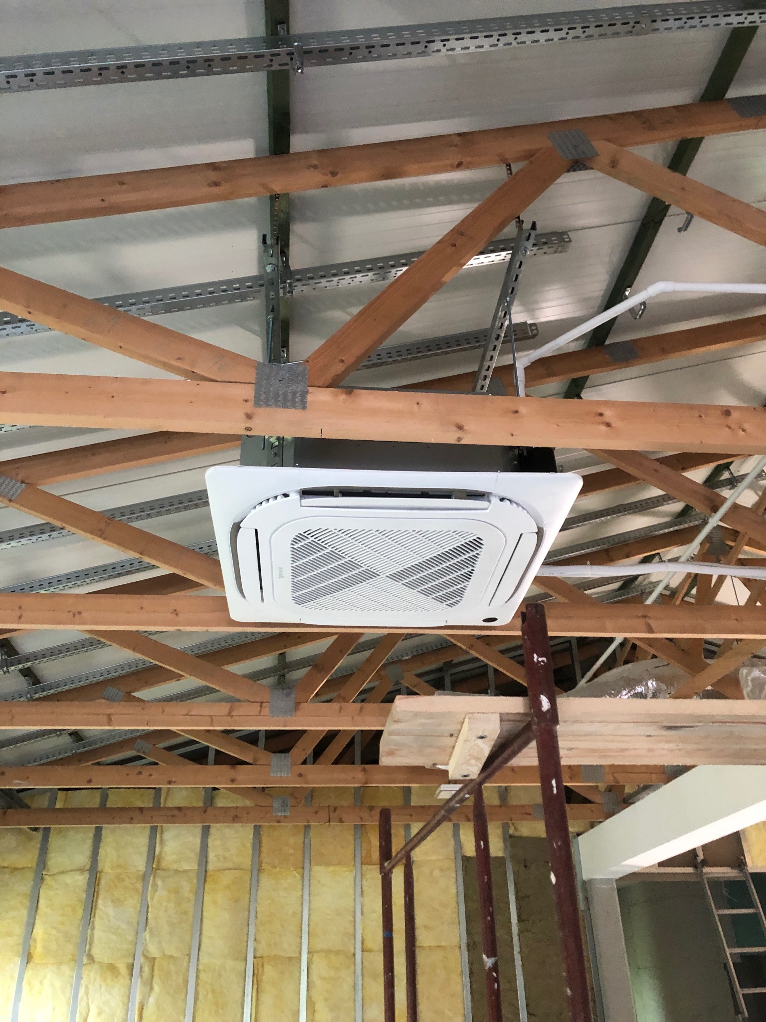 Biała kaseta klimatyzacyjna zamontowana pod drewnianą konstrukcją dachu, widoczne metalowe profile mocujące i ocieplenie ścian wełną mineralną.