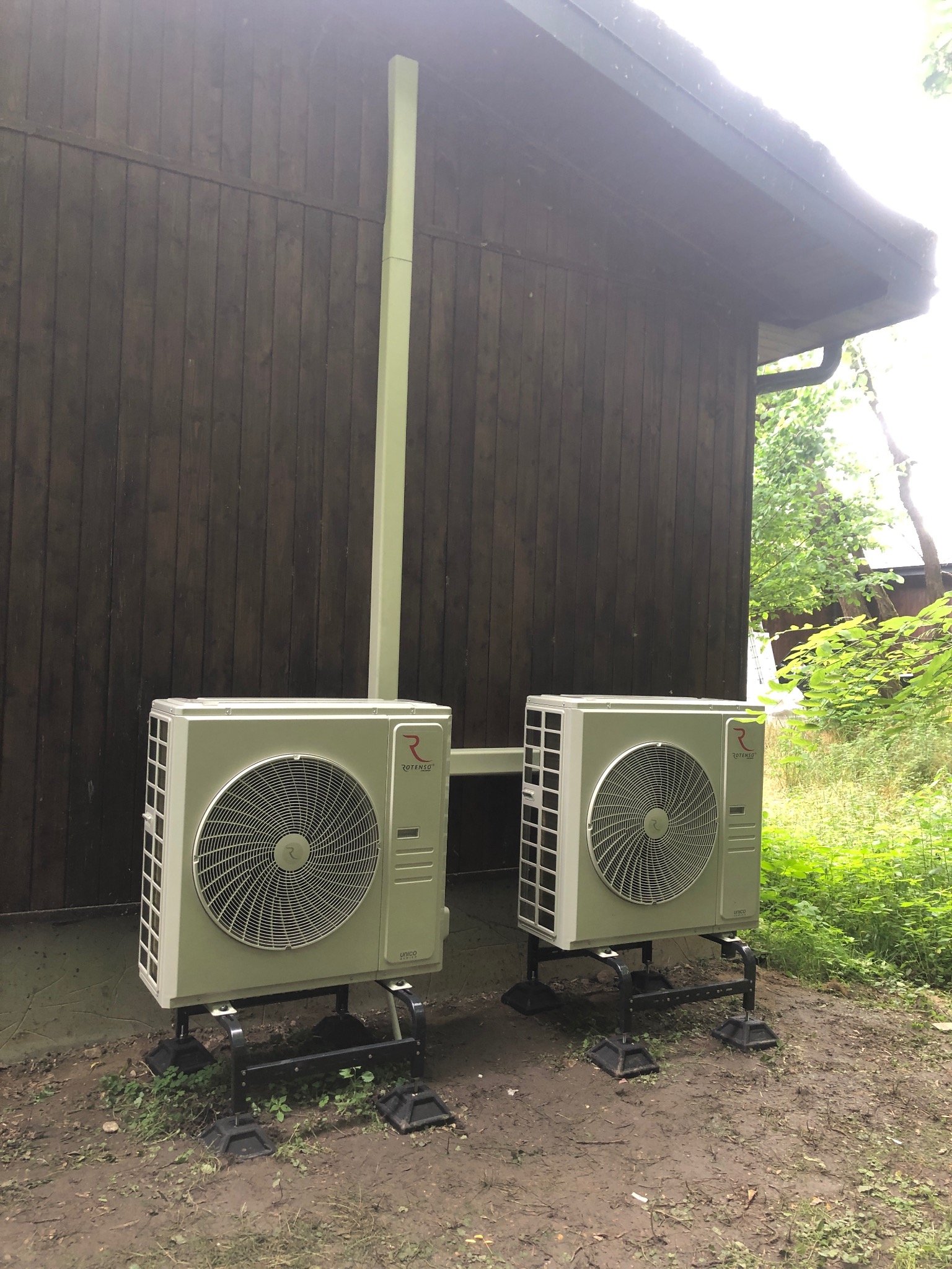 Dwa zewnętrzne agregaty klimatyzacji Rotenso zamontowane na czarnych podstawach na zewnątrz drewnianego budynku. Widoczne rury chłodnicze w kolorze jasnej zieleni.