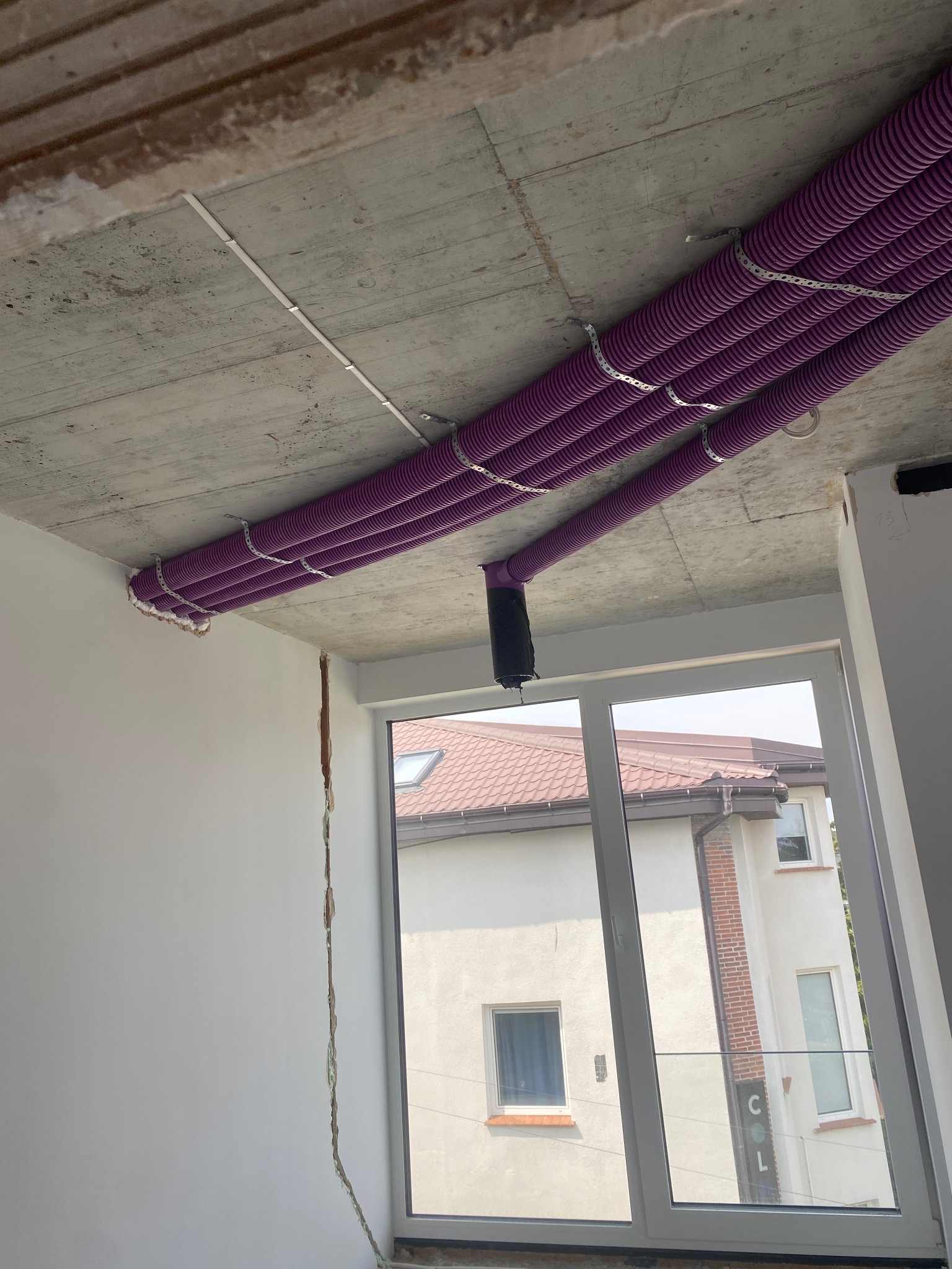 Fioletowe rury rekuperacji zamontowane pod surowym sufitem w budynku w Jesionce, widoczne przez okno sąsiednie budynki.