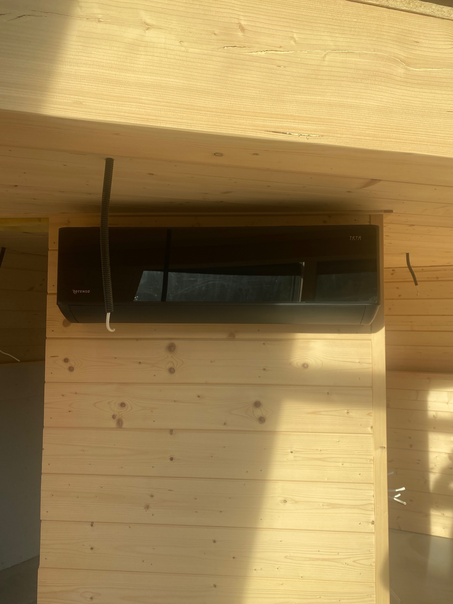 Czarna klimatyzacja Rotenso Teta zamontowana na drewnianej ścianie z boazerii, widoczne rury instalacyjne i surowe drewniane belki stropowe.