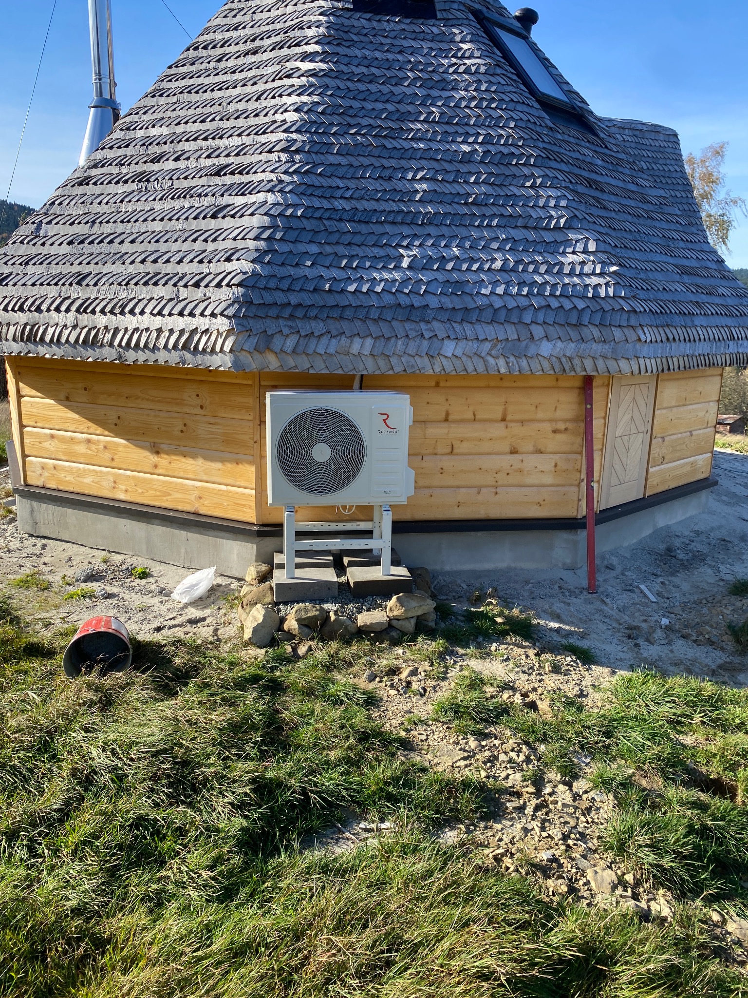 Zewnętrzna jednostka klimatyzacji zamontowana przy drewnianym budynku z dachem pokrytym gontem, widoczny poziomica i wiadro