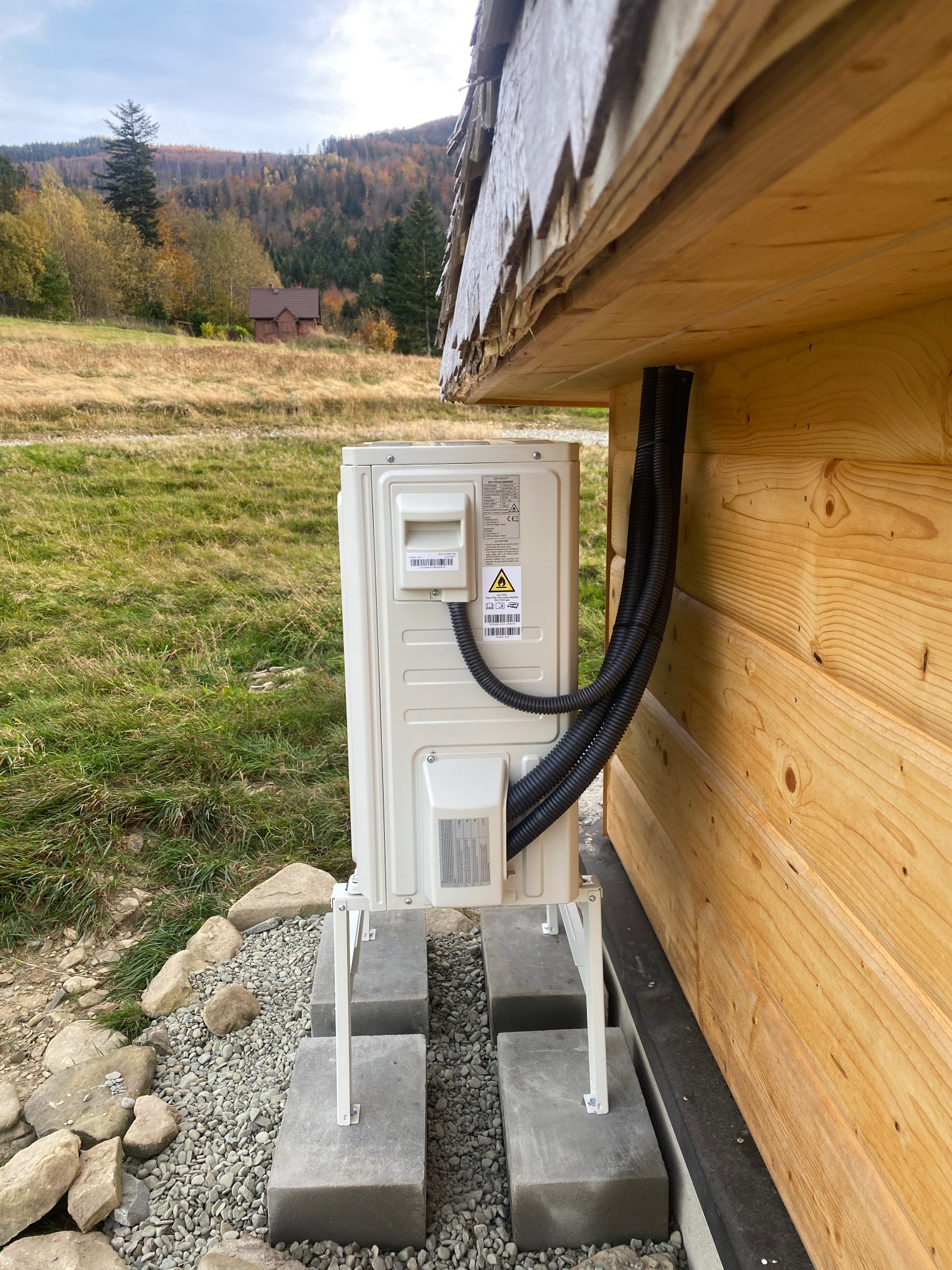 Zewnętrzna jednostka klimatyzacji zamontowana na betonowych podstawach przy drewnianym domu z widokiem na górski krajobraz.