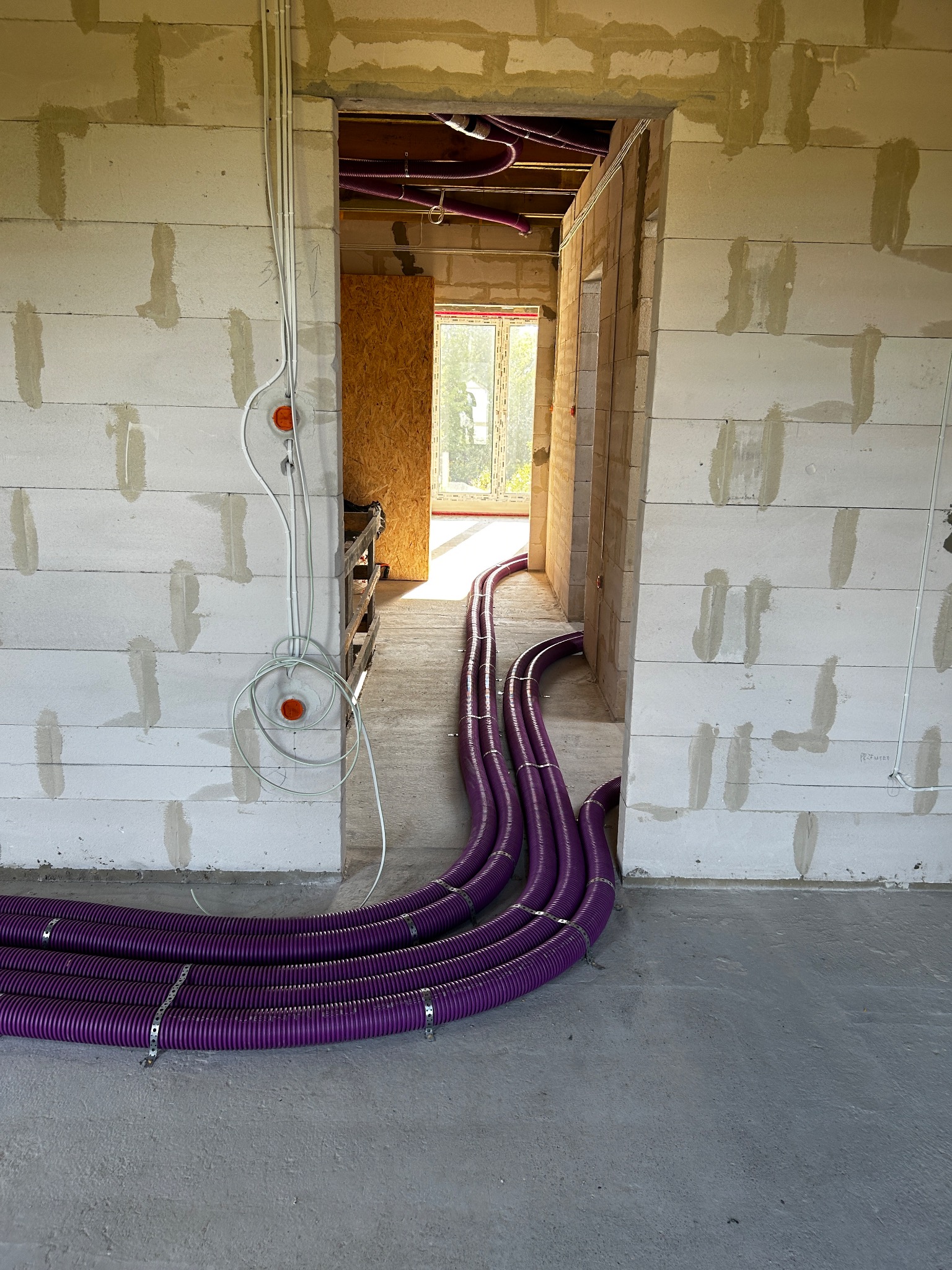 Fioletowe rury wentylacyjne prowadzone po podłodze w surowym wnętrzu budynku z białych bloczków, widoczne otwory drzwiowe i okno w tle.