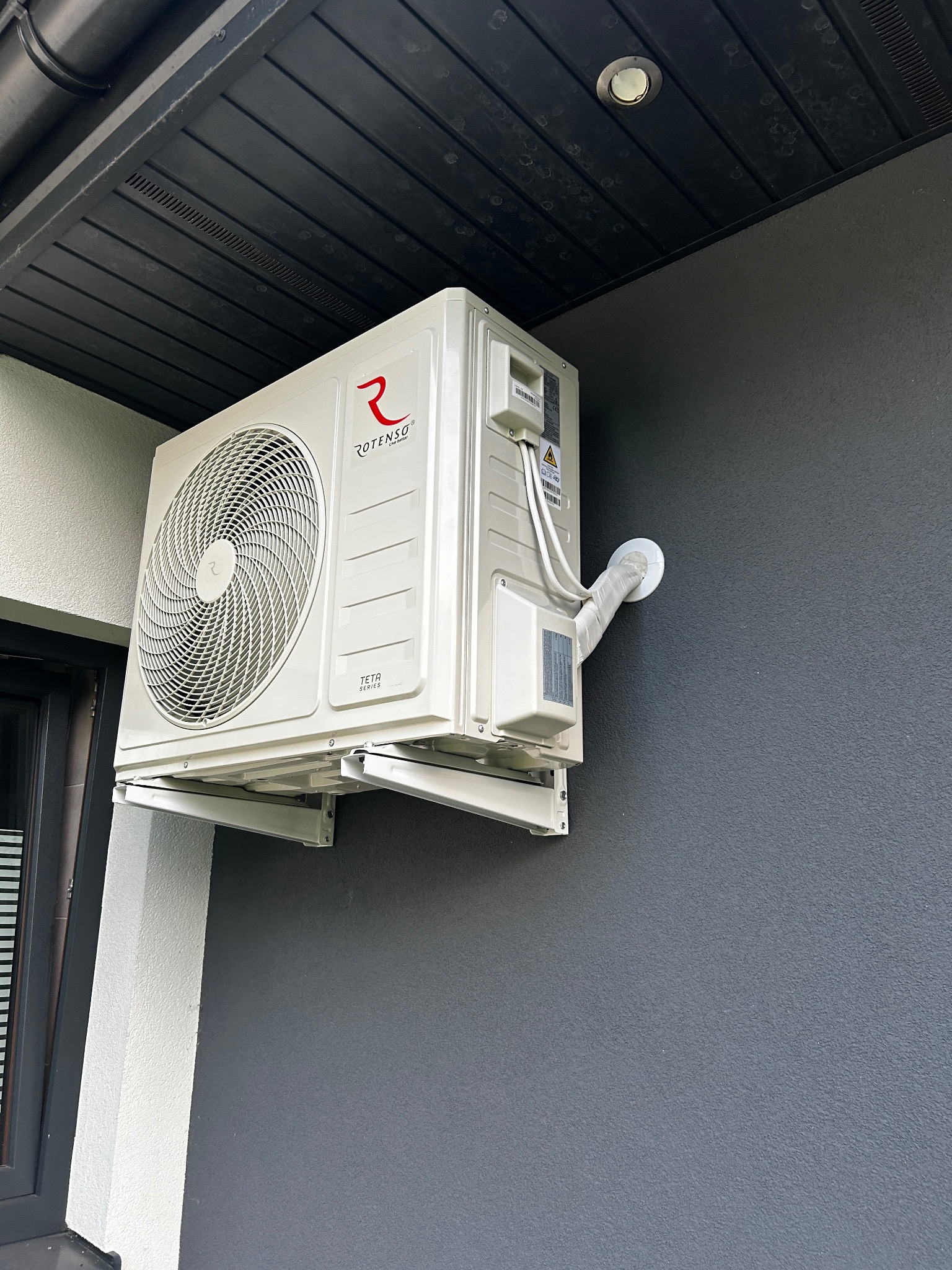 Biała jednostka zewnętrzna klimatyzatora Rotenso zamontowana na szarej ścianie budynku, widoczne przewody w izolacji i elementy montażowe.