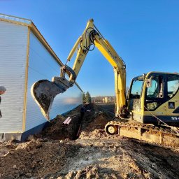 Szymon Czysz - Żółta koparka Hyundai wykonuje wykop pod rury wzdłuż ściany budynku. Widoczny pracownik w oddali i ułożone rury w wykopie. Błękitne niebo w tle.