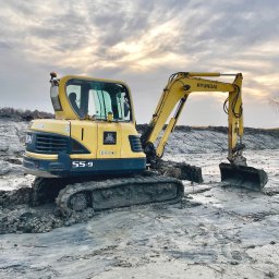 Szymon Czysz - Żółta mini koparka Hyundai Robex 55-9 na błotnistym terenie, z widocznym logiem GR Czyszcz. Pochmurne niebo w tle, sugerujące prace ziemne w trudnych warunkach.