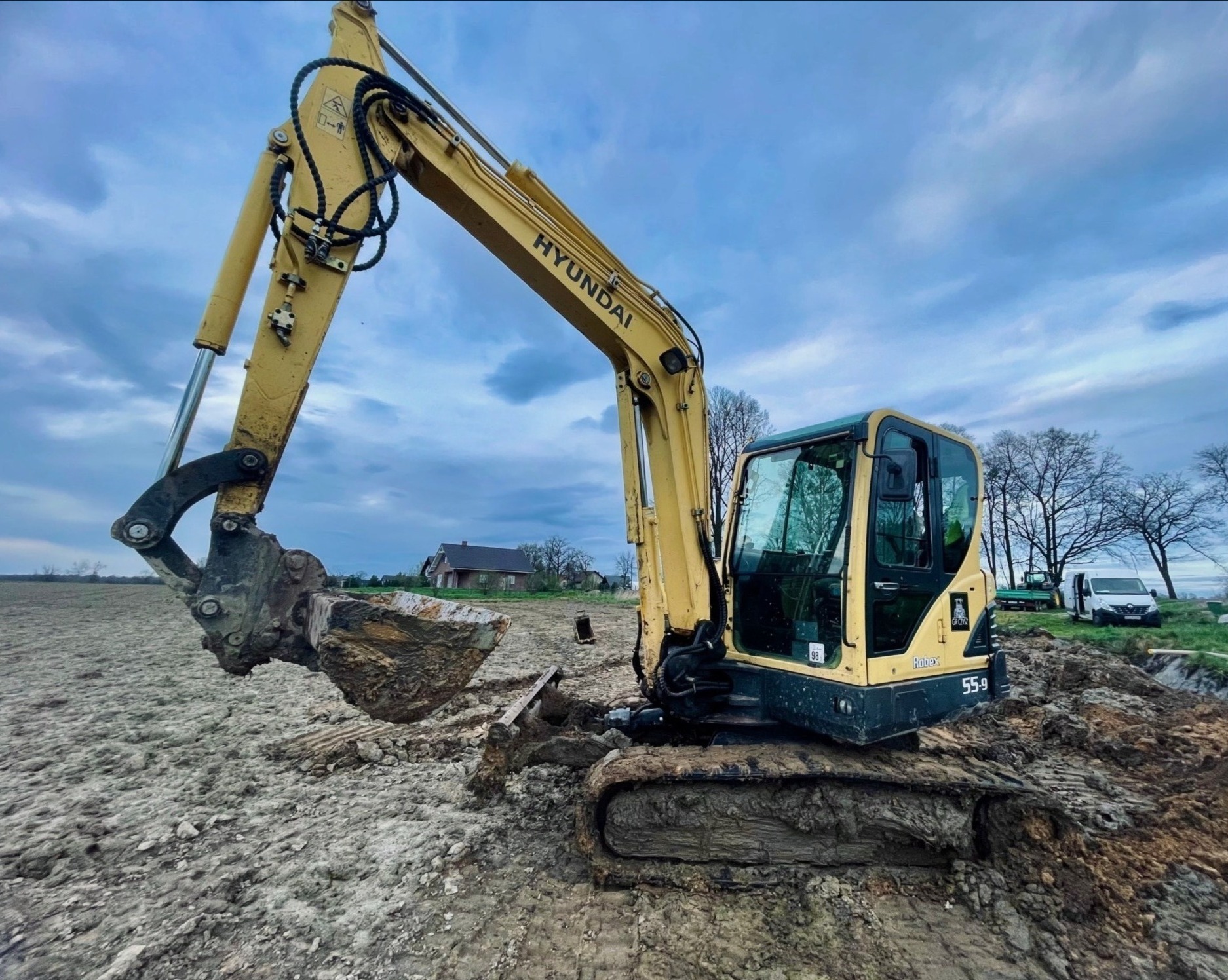 Żółta koparka Hyundai na polu, z łyżką wypełnioną ziemią, w tle budynek gospodarczy i pojazdy, pochmurne niebo.