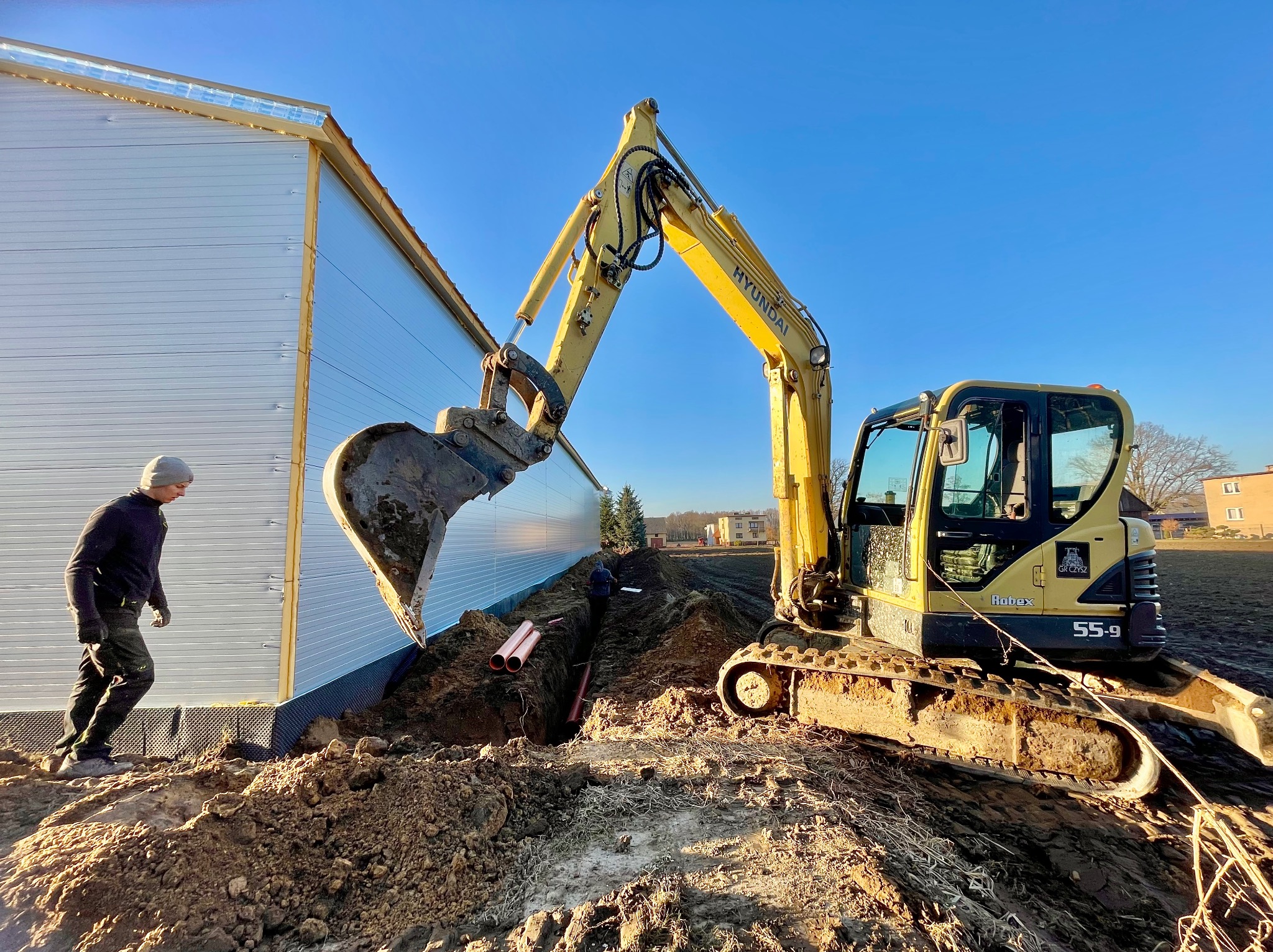Żółta koparka Hyundai wykonuje wykop pod rury wzdłuż ściany budynku. Widoczny pracownik w oddali i ułożone rury w wykopie. Błękitne niebo w tle.