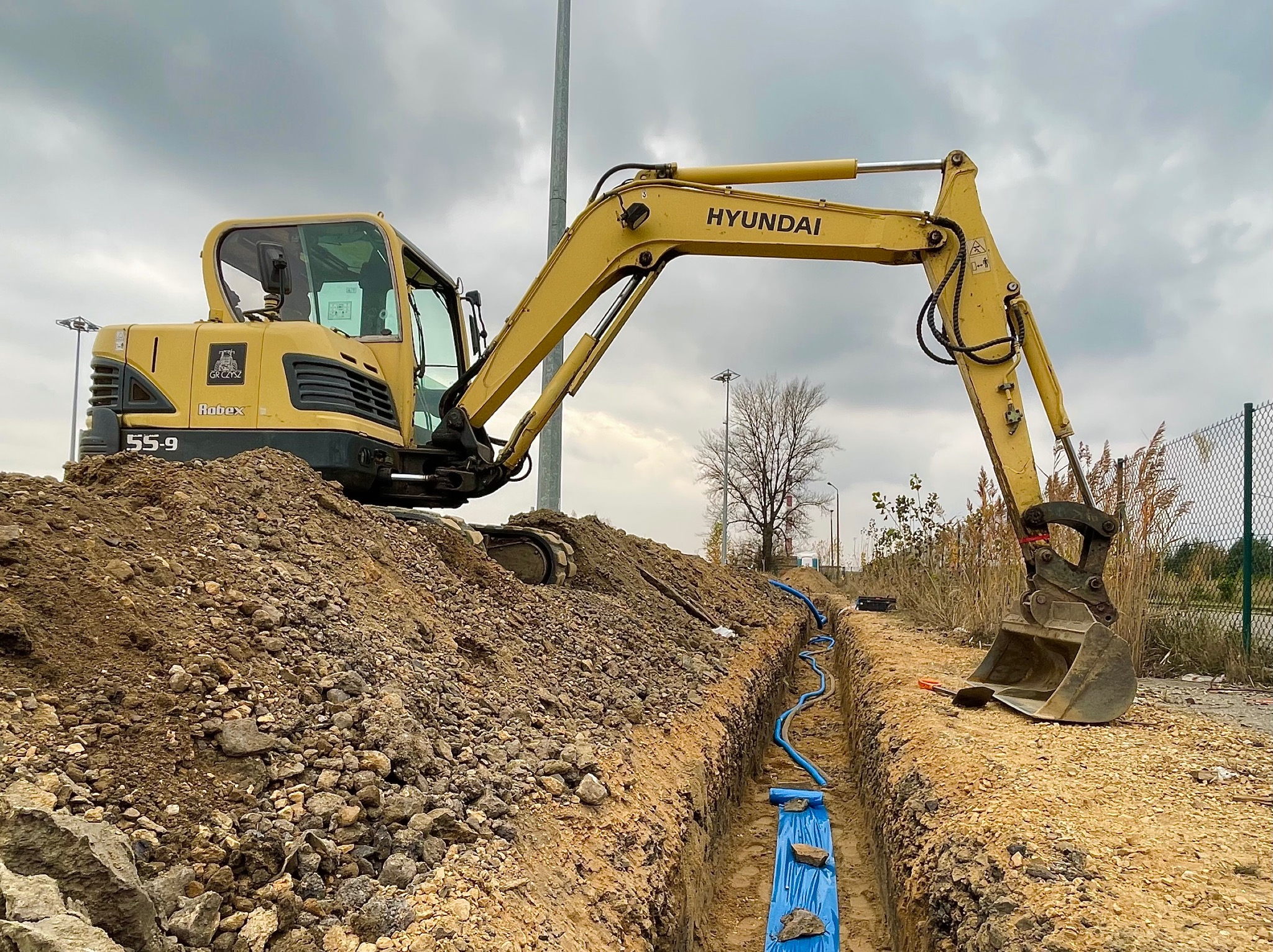 Żółta koparka Hyundai Robex 55-9 na placu budowy, wykonująca wykop pod niebieskie rury, zabezpieczone folią. Widoczne narzędzia i ziemia.