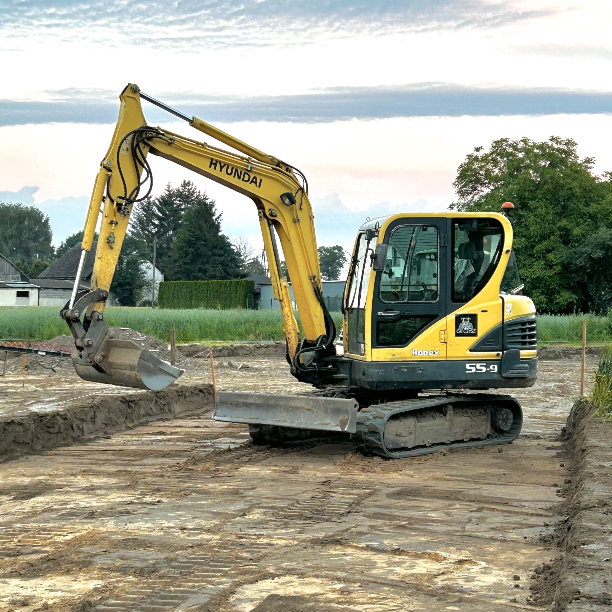 Żółta koparka Hyundai Robex 55-9 na placu budowy, w trakcie wykonywania wykopu pod fundamenty. Widoczne ślady opon na ziemi i wykopane rowy.