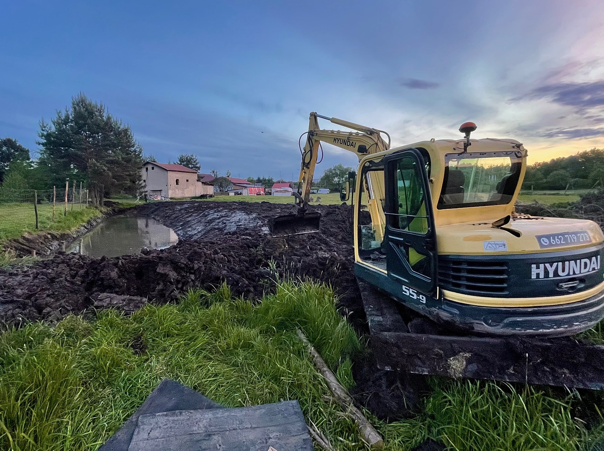 Żółta koparka Hyundai 55-9 w trakcie pogłębiania stawu na tle wiejskiego krajobrazu o zmierzchu. Widoczne świeżo wykopane błoto i zielona trawa.