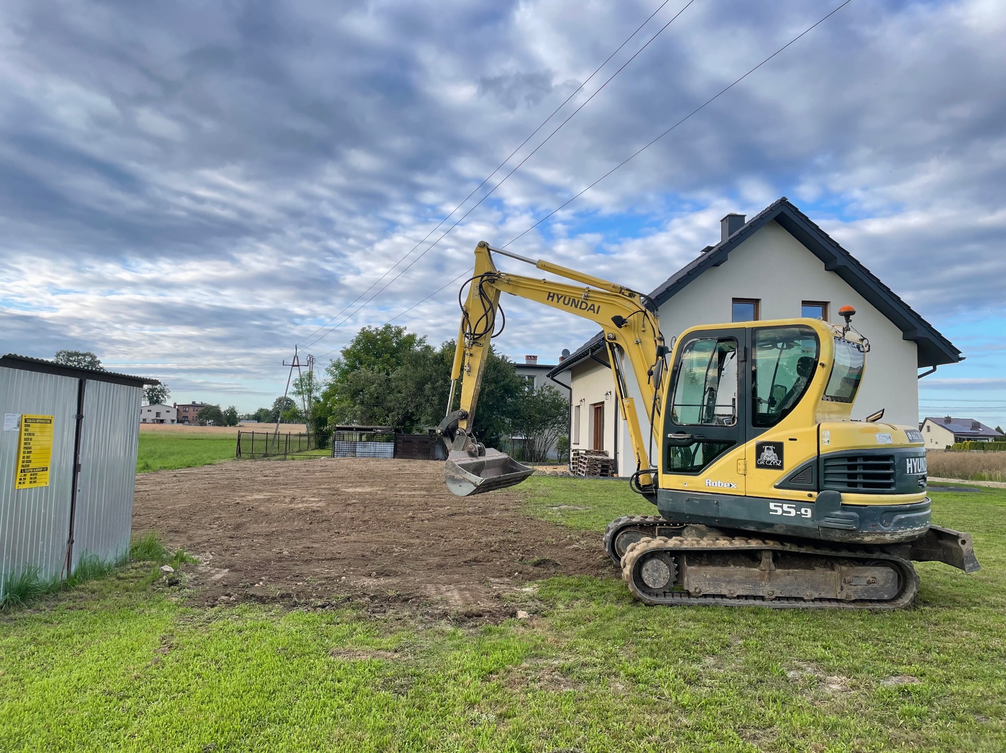 Żółta koparka Hyundai 55-9 na działce w Mizerowie po wykonaniu prac ziemnych. W tle dom i pochmurne niebo. Widoczny wykop pod przyszłą budowę.