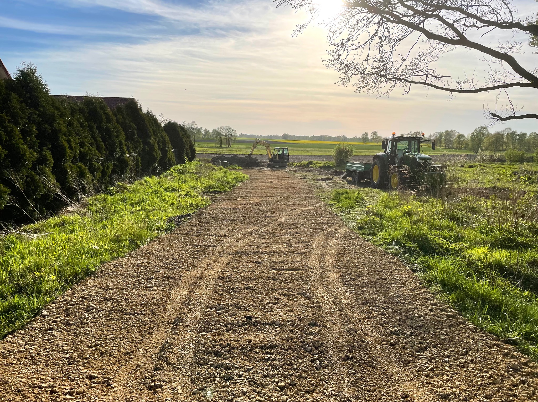 Utwardzona droga gruntowa z widocznym traktorem i koparką w tle, otoczona zielenią i drzewami, pod błękitnym niebem z promieniami słońca.