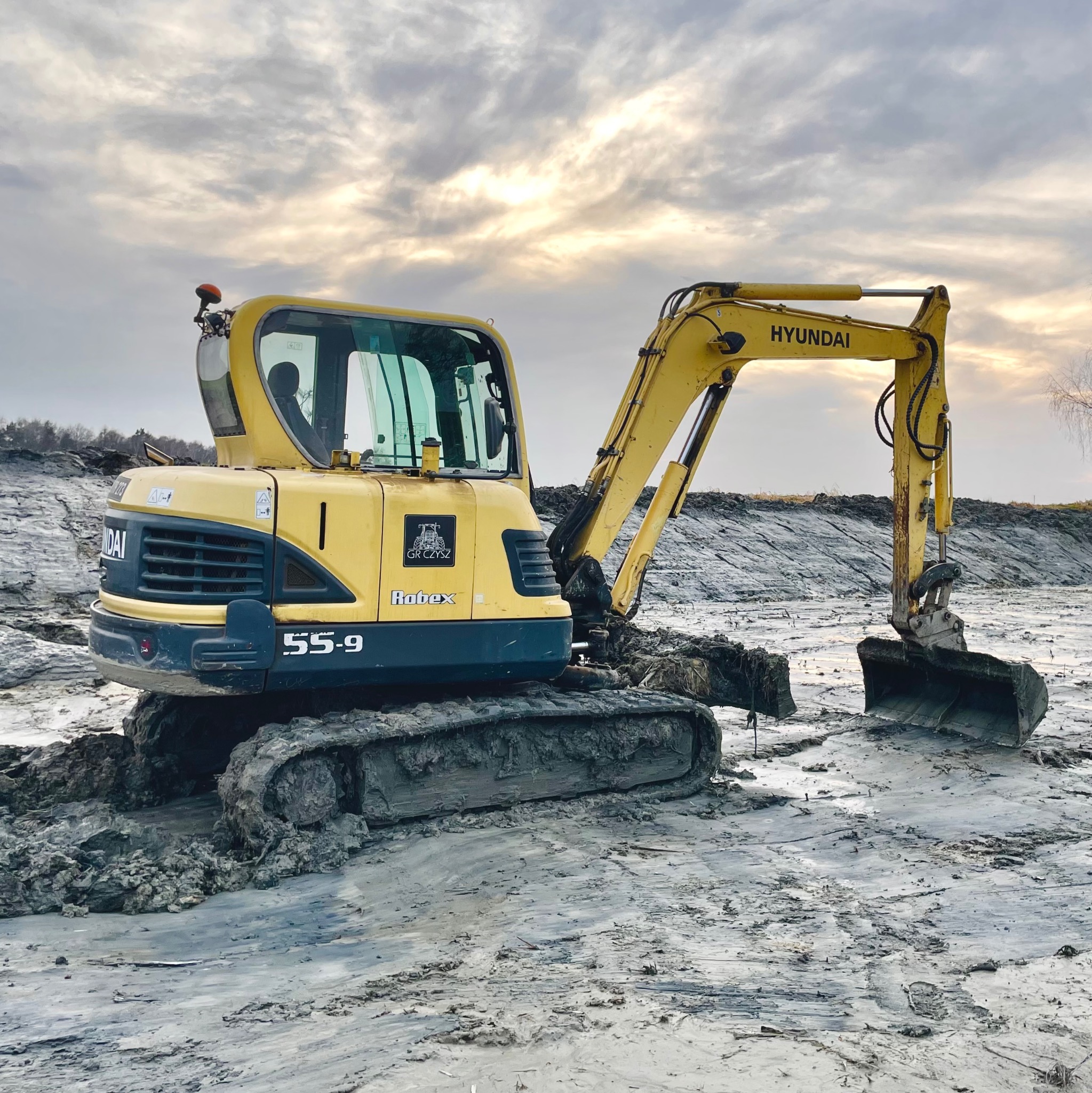 Żółta mini koparka Hyundai Robex 55-9 na błotnistym terenie, z widocznym logiem GR Czyszcz. Pochmurne niebo w tle, sugerujące prace ziemne w trudnych warunkach.