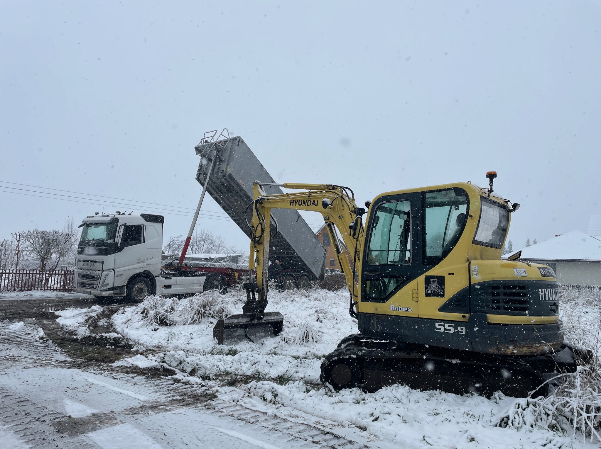 Żółta koparka Hyundai i biała ciężarówka wywrotka podczas załadunku ziemi w zimowy dzień. Śnieg pada, a ziemia jest błotnista.