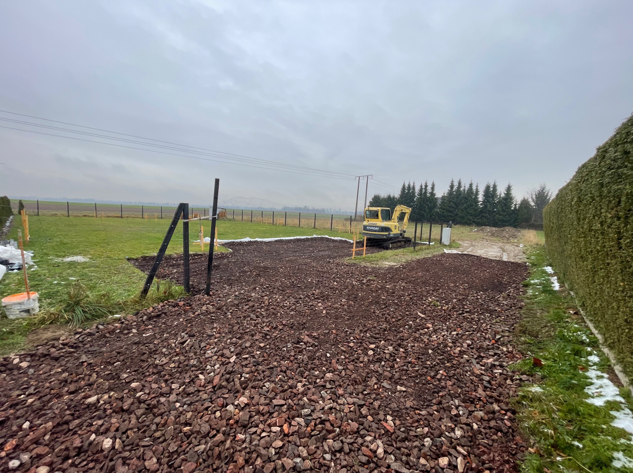 Żwirowa nawierzchnia z żółtą mini koparką Hyundai na działce, widoczne słupki ogrodzeniowe i zieleń. Pochmurne niebo.