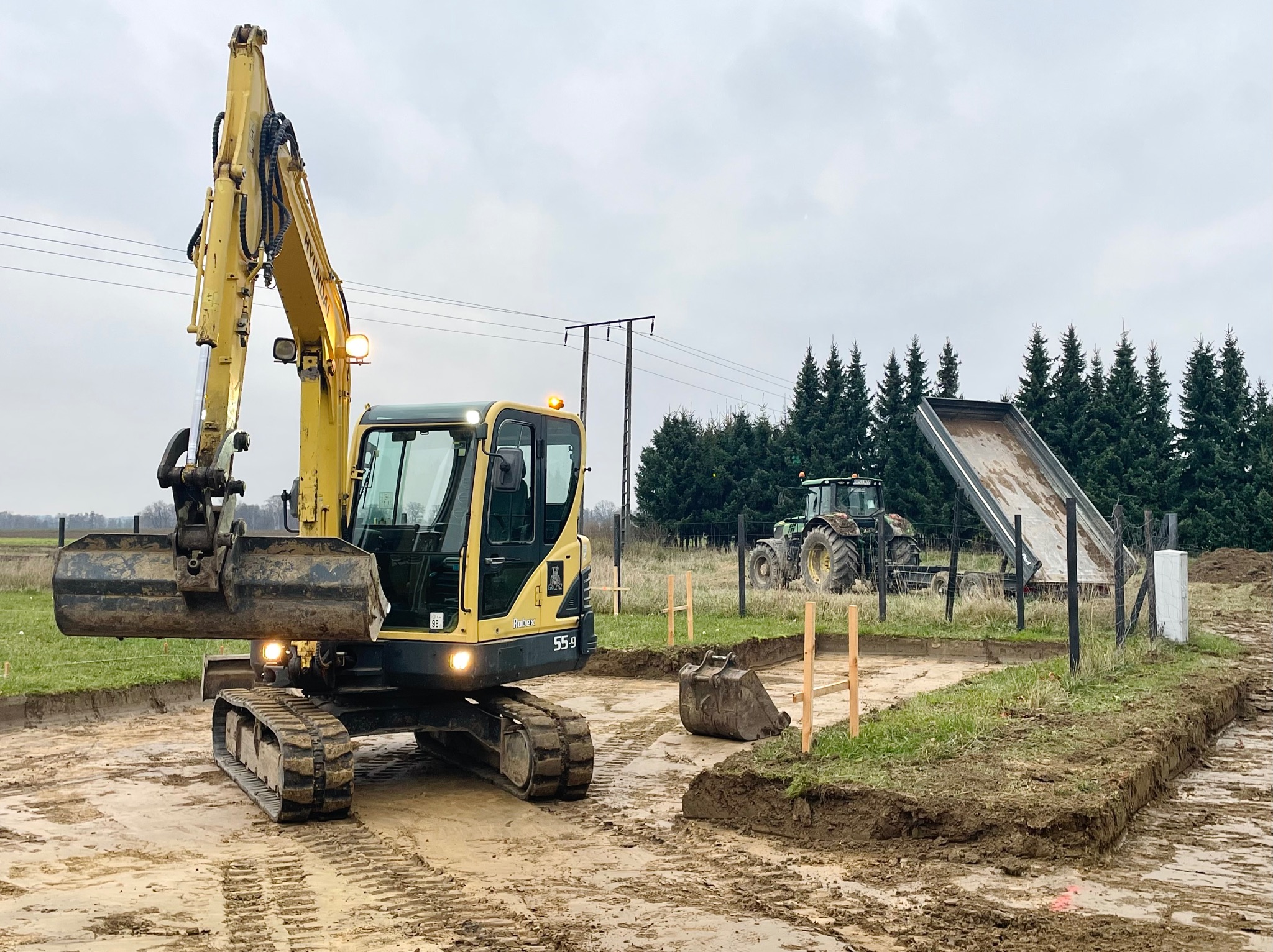 Żółta koparka Hyundai Robex 55-9 na wykopie pod fundamenty, w tle traktor z przyczepą i las iglasty. Widoczne ślady gąsienic i wytyczone granice wykopu.
