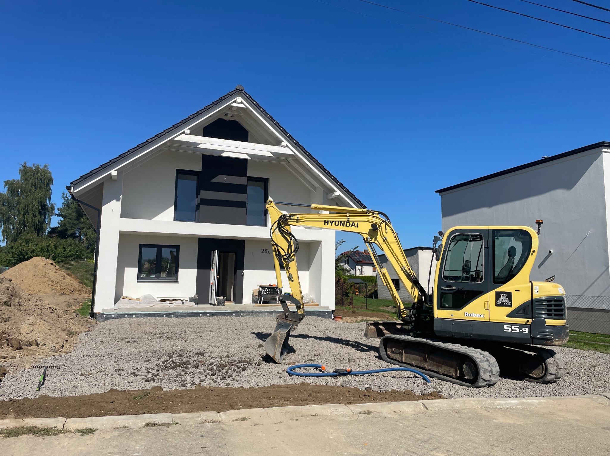 Żółta koparka Hyundai na żwirowym podłożu przed nowoczesnym, białym domem z czarnymi akcentami. W tle błękitne niebo i fragmenty zieleni. Etap przygotowania terenu.