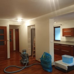 Jacek Bałuszyński LeoColor Usługi Malarsko-Wykończeniowe - Wnętrze po remoncie: świeżo pomalowane ściany w odcieniach bieli, drewniana podłoga, sprzęt do sprzątania i zabezpieczenia. Widok na kuchnię i korytarz.