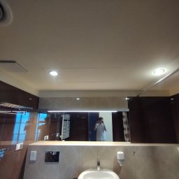 Jacek Bałuszyński LeoColor Usługi Malarsko-Wykończeniowe - Wykończona łazienka z brązowymi płytkami, białą umywalką i dużym lustrem, w którym odbija się osoba robiąca zdjęcie. Widoczne oświetlenie sufitowe i akcesoria łazienkowe.