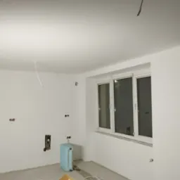 Białe ściany po malowaniu w pokoju z oknem i grzejnikiem. Widoczne gniazdka elektryczne i przewody. Świeżo pomalowane wnętrze w trakcie wykańczania.