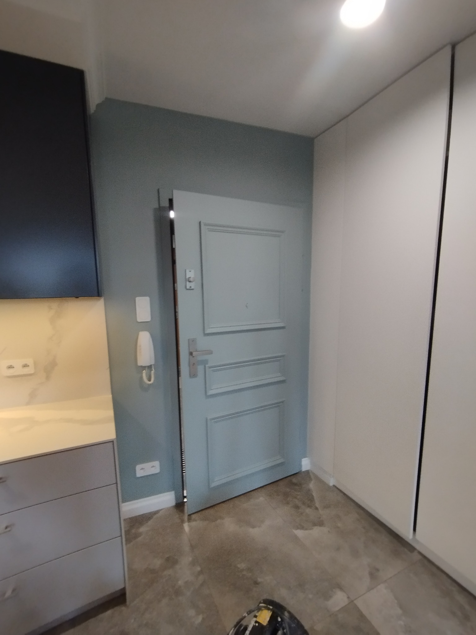 Błękitne drzwi wewnętrzne z panelami w odnowionym mieszkaniu, widoczne z perspektywy podłogi z szarych płytek. Ściana w kolorze gołębim, biała szafa w zabudowie.