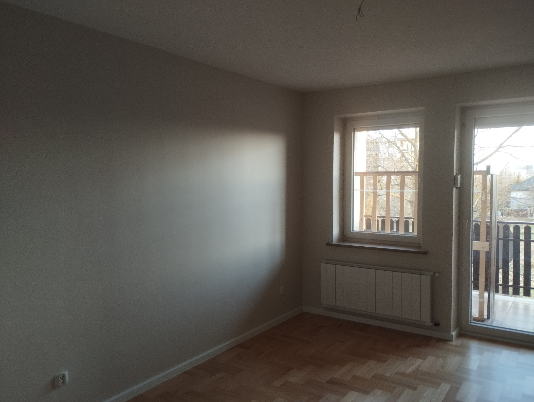 Jasne, świeżo pomalowane wnętrze pokoju z parkietem, oknem i wyjściem na balkon. Ściany w odcieniach beżu, widoczny grzejnik. Minimalistyczny wystrój.