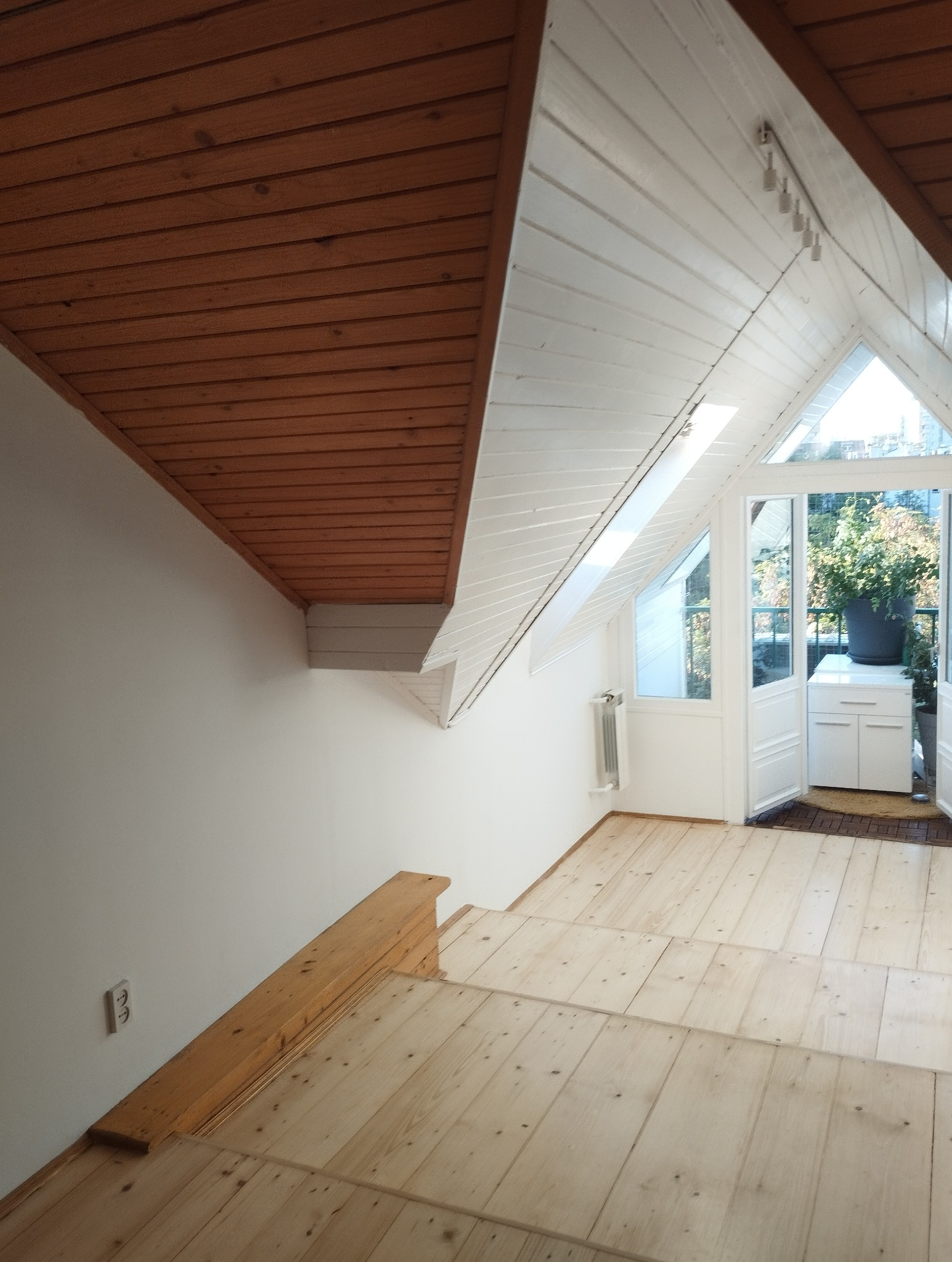 Jasne, drewniane schody wiodą do przeszklonego pokoju z roślinami. Białe ściany kontrastują z brązowym, drewnianym sufitem. Widoczny grzejnik i gniazdko elektryczne.