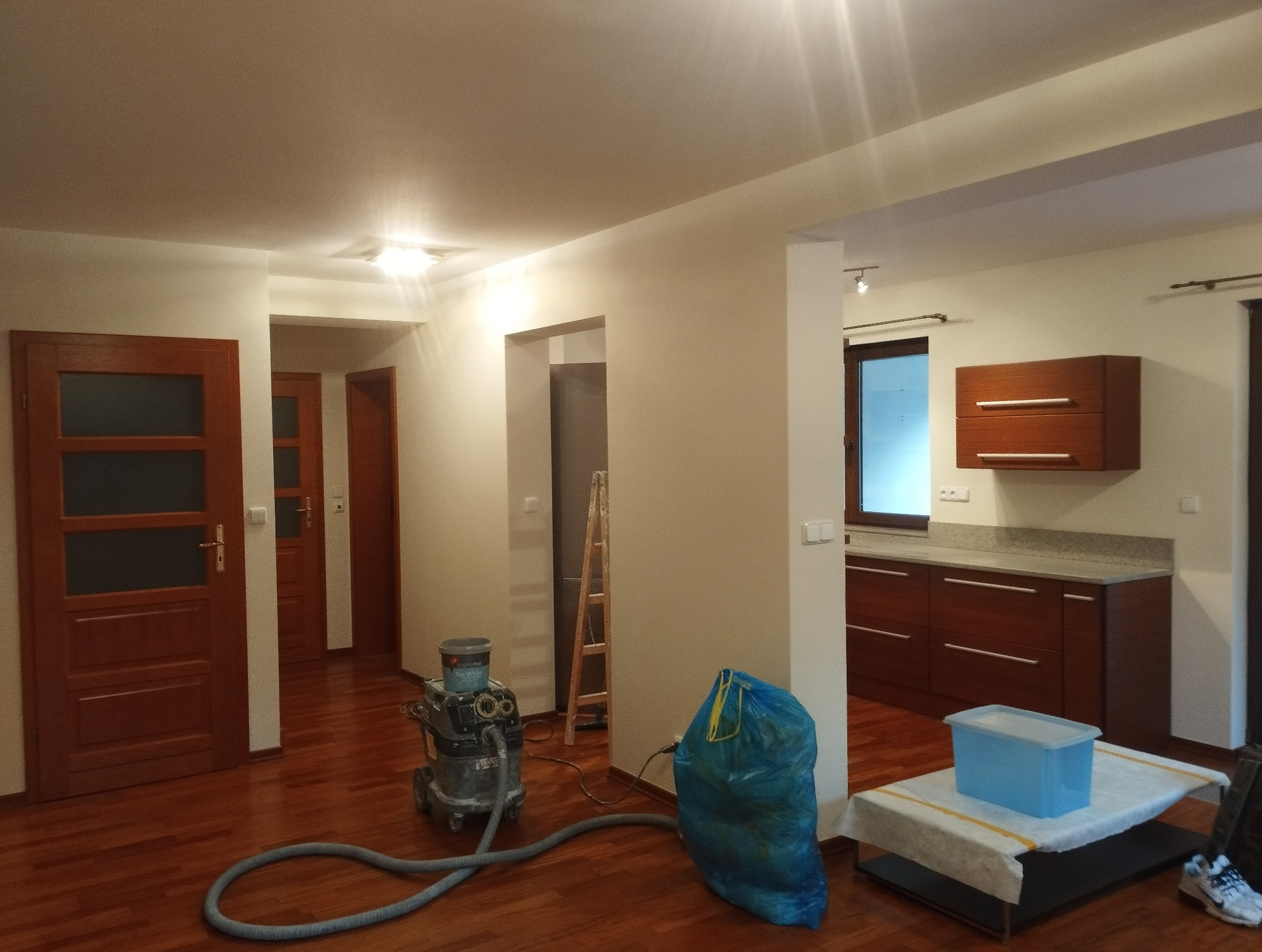Wnętrze po remoncie: świeżo pomalowane ściany w odcieniach bieli, drewniana podłoga, sprzęt do sprzątania i zabezpieczenia. Widok na kuchnię i korytarz.