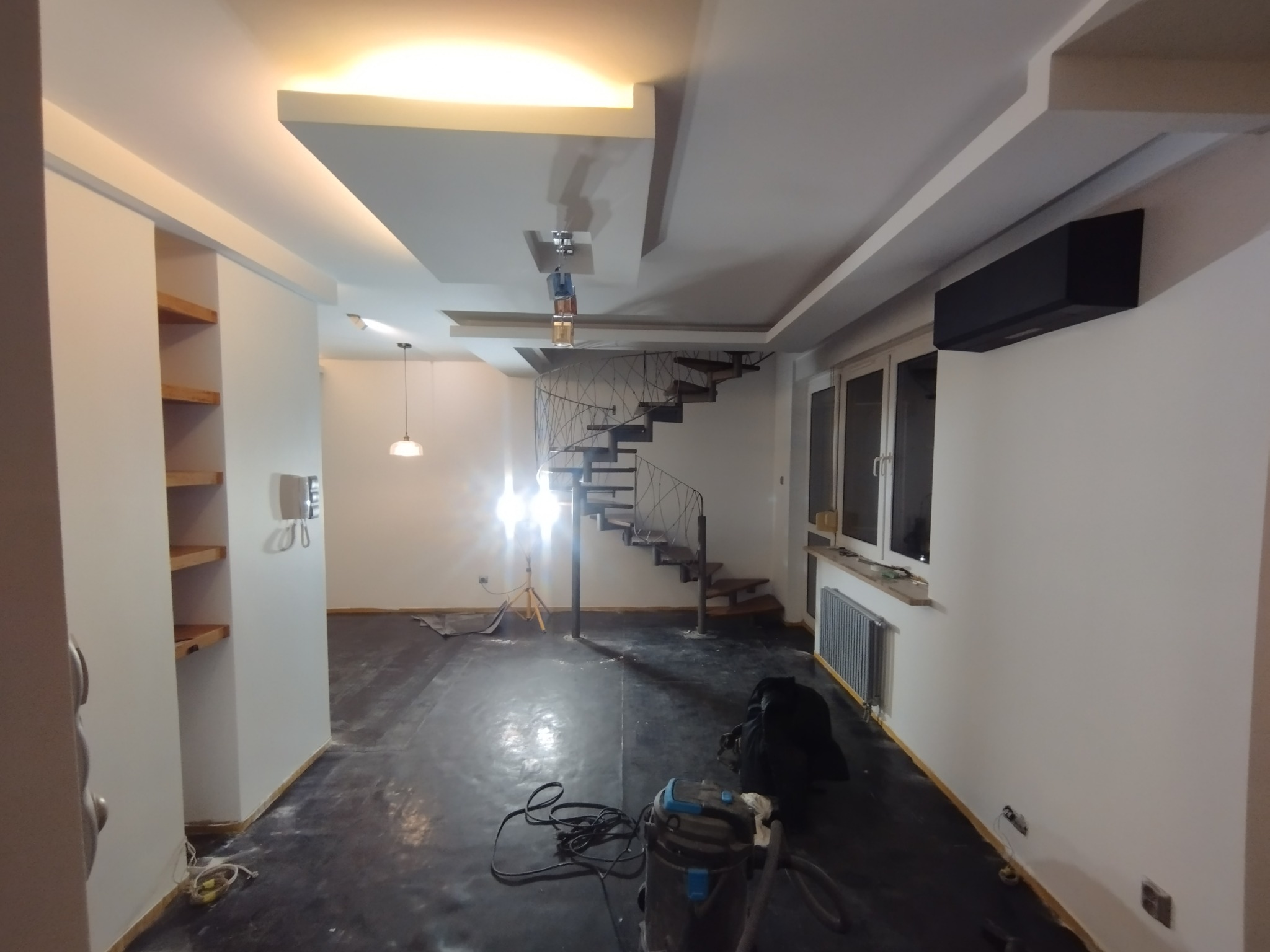 Wnętrze po remoncie: białe ściany, spiralne schody, zabezpieczona podłoga, widoczny odkurzacz przemysłowy. Oświetlenie sufitowe i lampa wisząca.