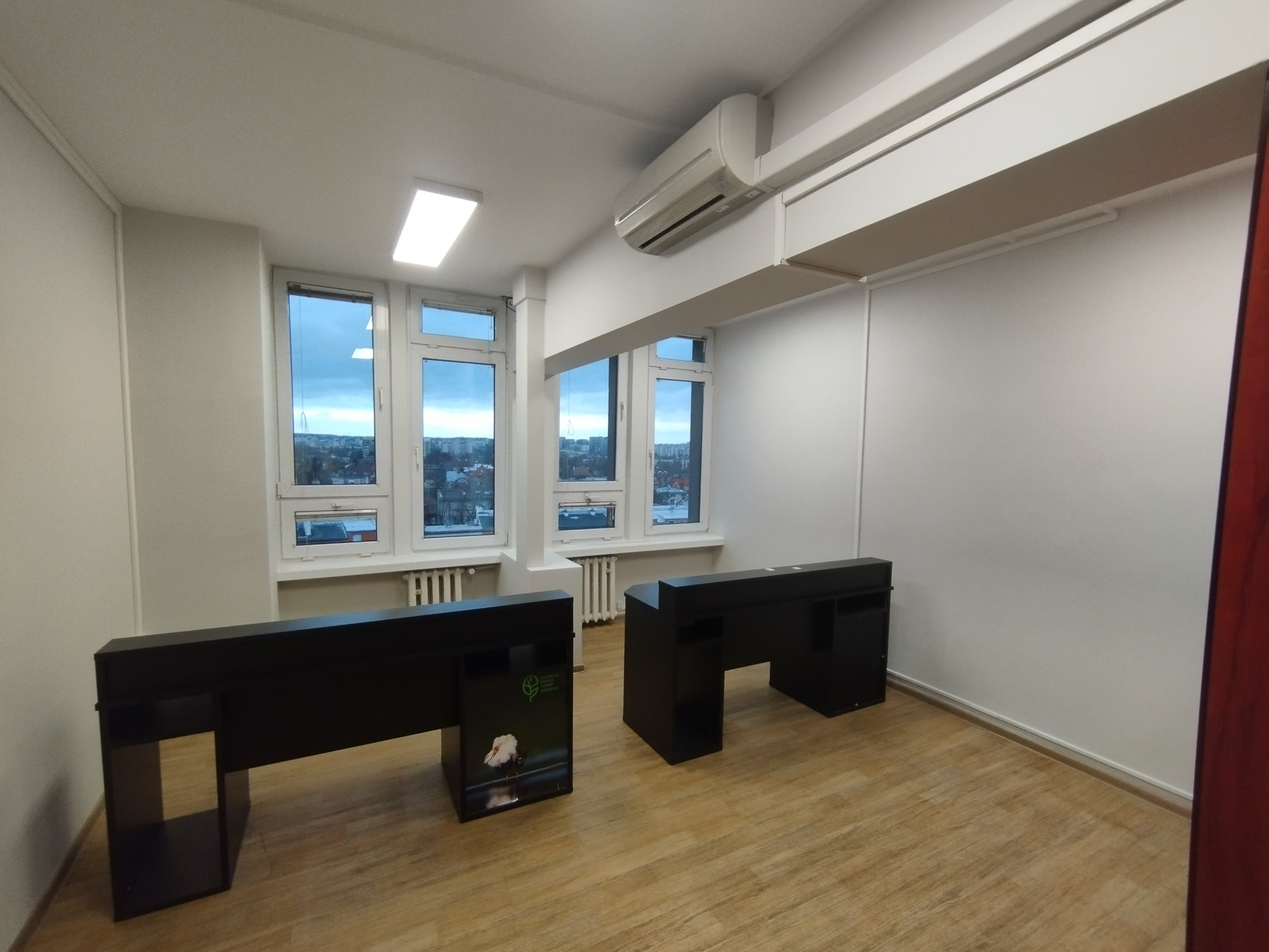 Jasne biuro po malowaniu: białe ściany, drewniana podłoga, czarne biurka, okna z widokiem na miasto. Minimalistyczny design, czystość i porządek.