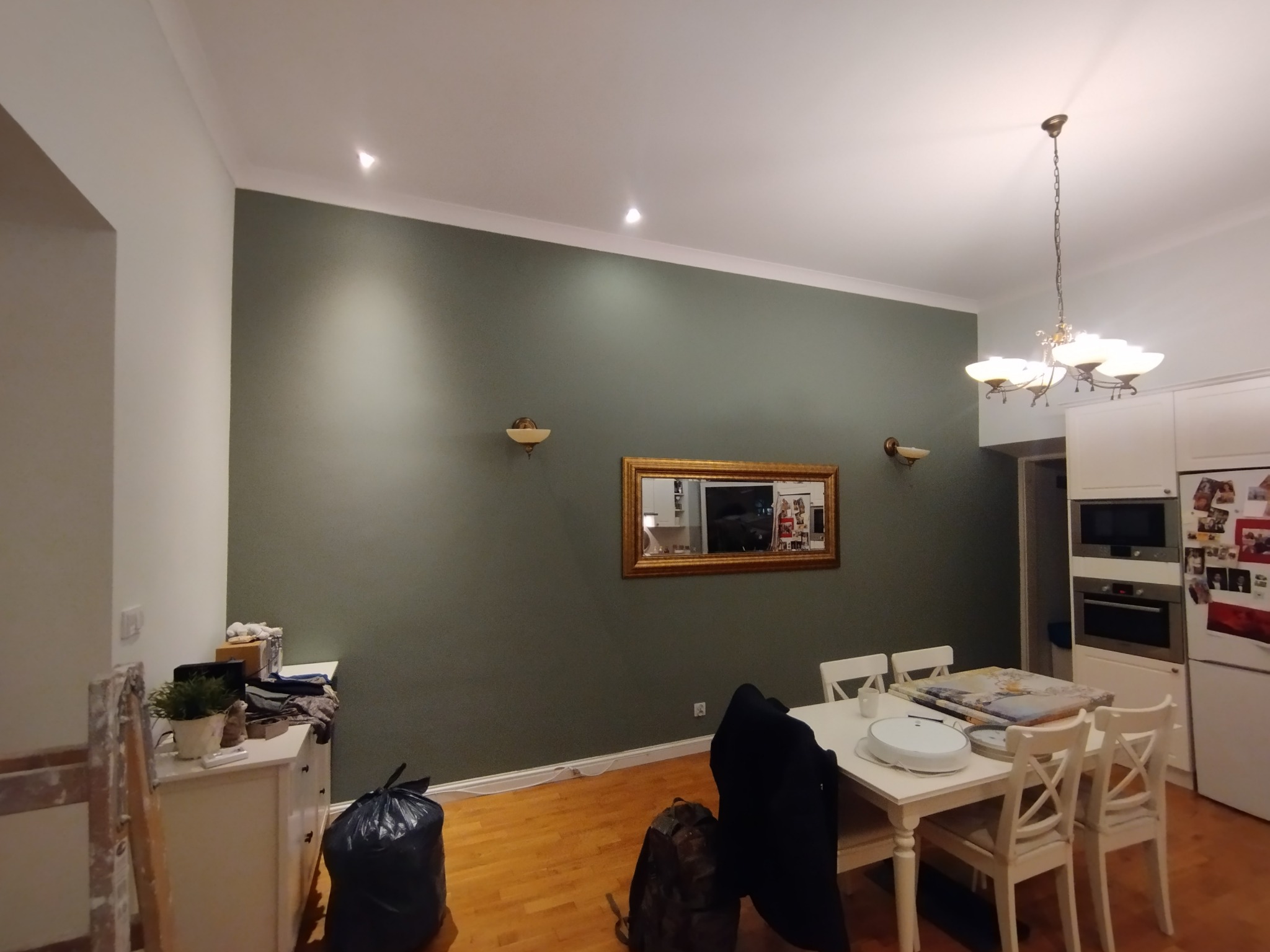 Ściana w odcieniach zieleni z dekoracyjnym lustrem w złotej ramie, oświetlona kinkietami. Widoczny stół z krzesłami i elementy wyposażenia wnętrza.