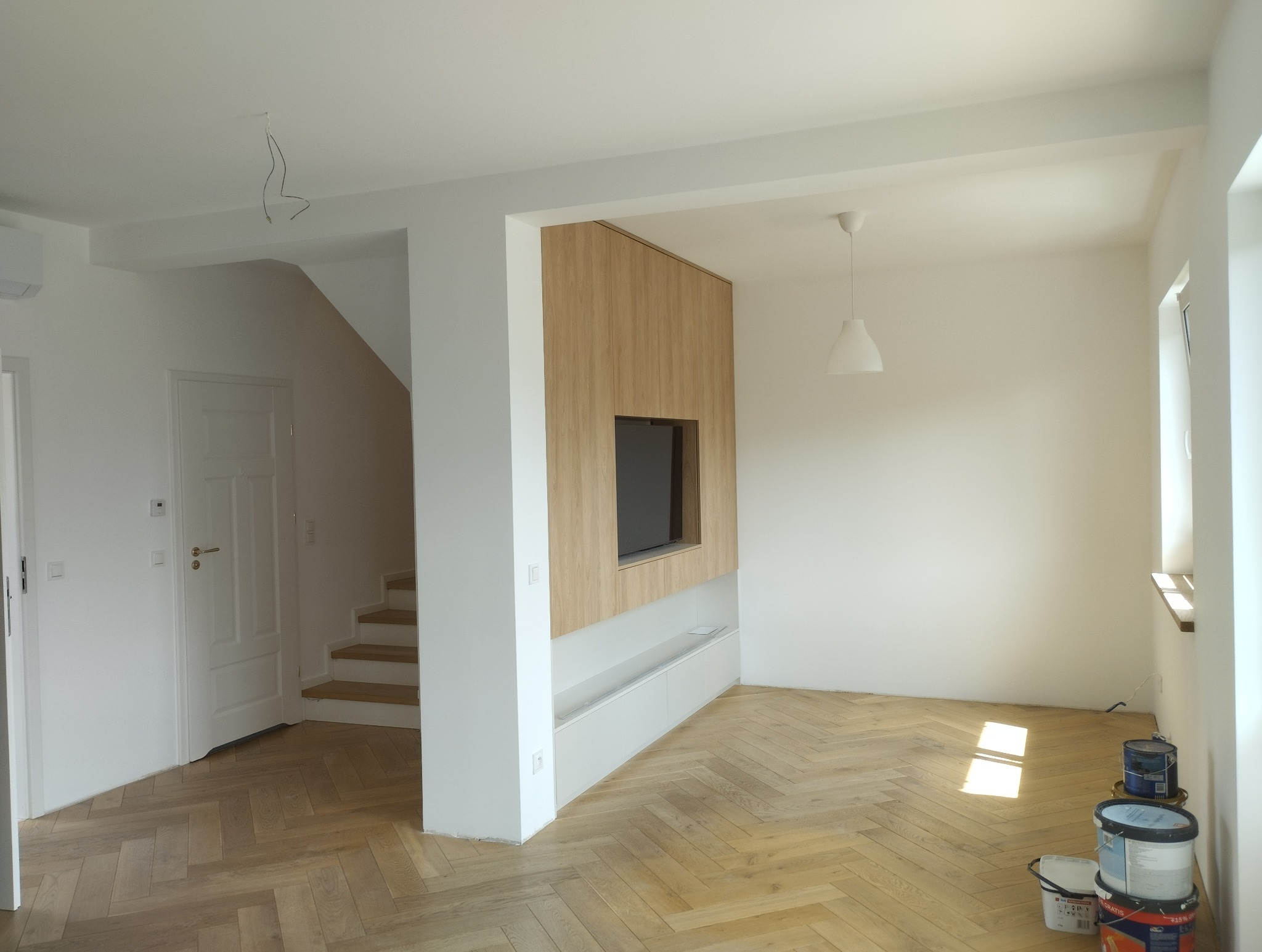 Jasne wnętrze po remoncie: świeżo pomalowane ściany, parkiet w jodełkę, zabudowa TV z drewnianym panelem. Widoczne puszki z farbą.