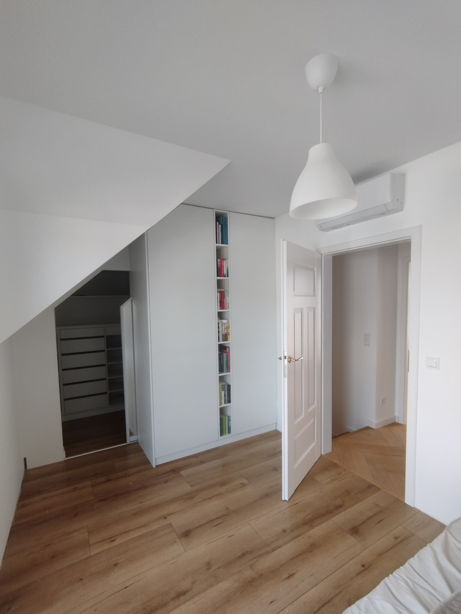 Jasne wnętrze pokoju na poddaszu z drewnianą podłogą, białymi ścianami i szafą wnękową z półkami na książki. Widoczny fragment łóżka i otwarte drzwi do kolejnego pomieszczenia.