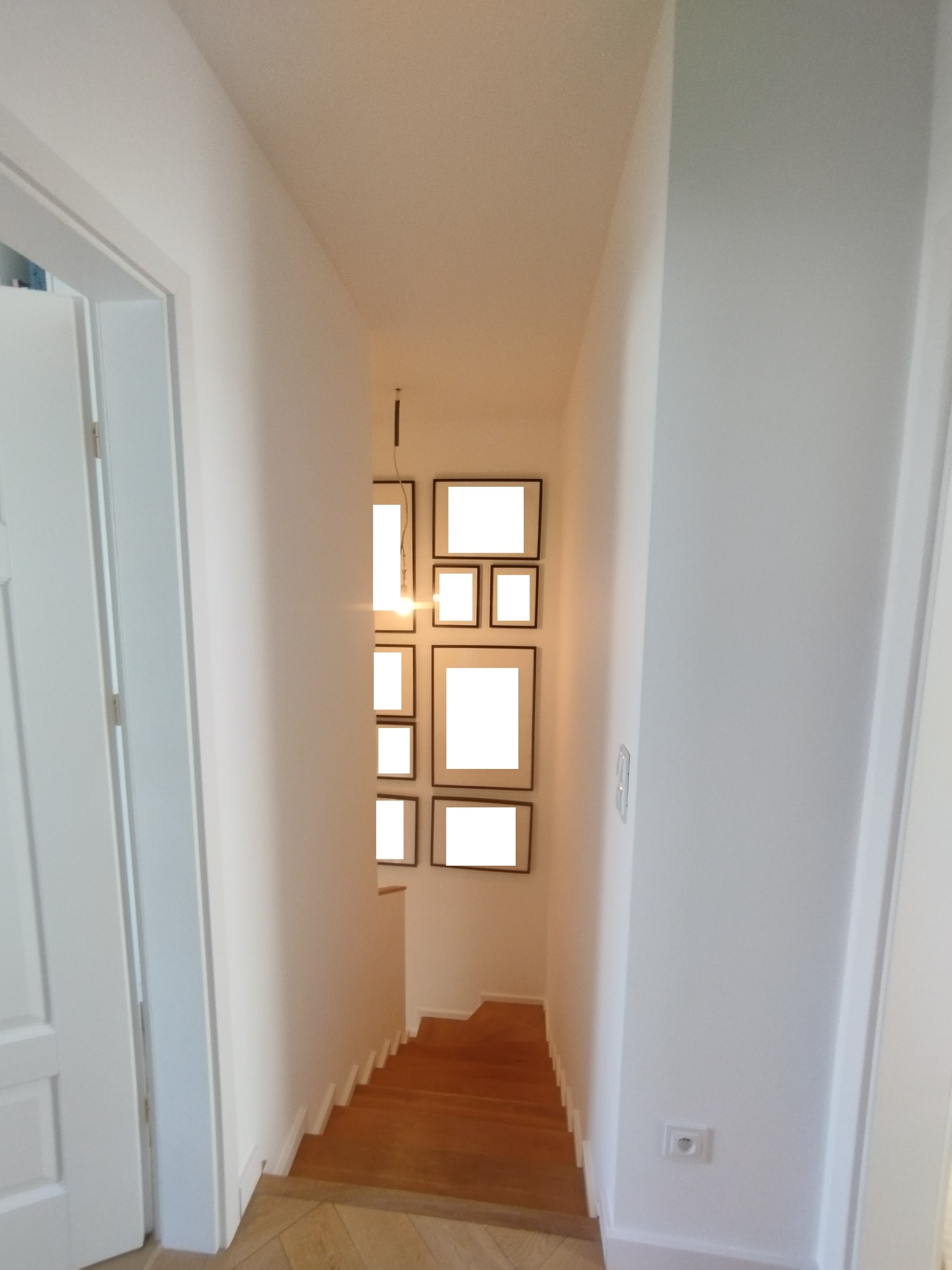 Białe ściany korytarza z drewnianymi schodami i galerią pustych ramek na ścianie, widok z perspektywy drzwi. Minimalistyczny design.