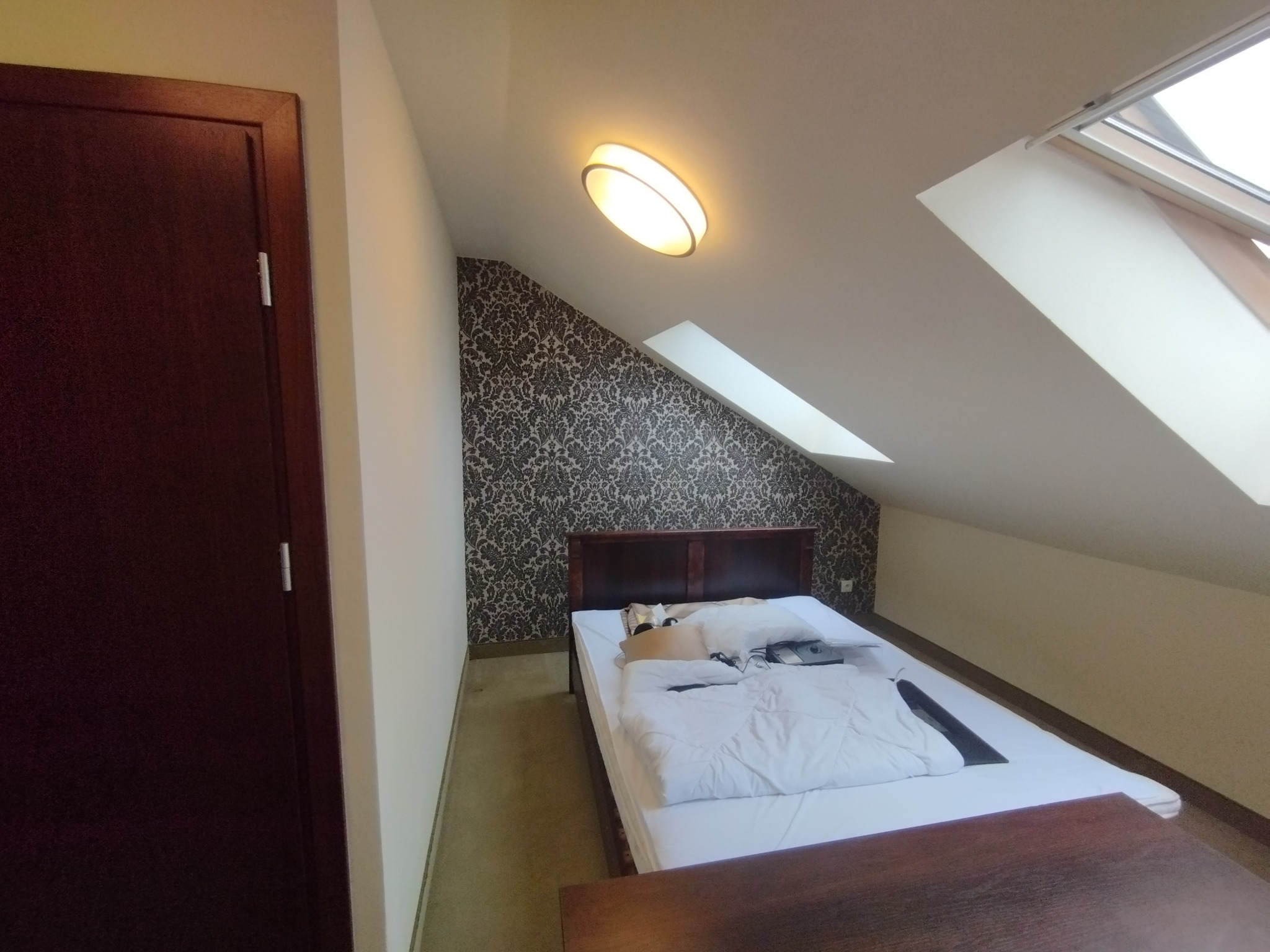 Pokój na poddaszu z łóżkiem, wzorzystą tapetą i oknem dachowym. Widoczna lampa sufitowa i ciemne drzwi po lewej stronie. Ujęcie z dołu, podkreślające skosy.