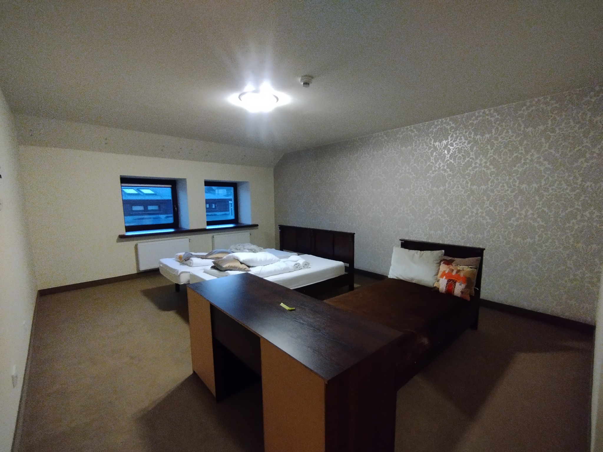 Pokój hotelowy po odświeżeniu: dwie szafki nocne, dwa łóżka z pościelą, ściana z wzorzystą tapetą, dywan na podłodze. Widok na okna z widokiem na zewnątrz.