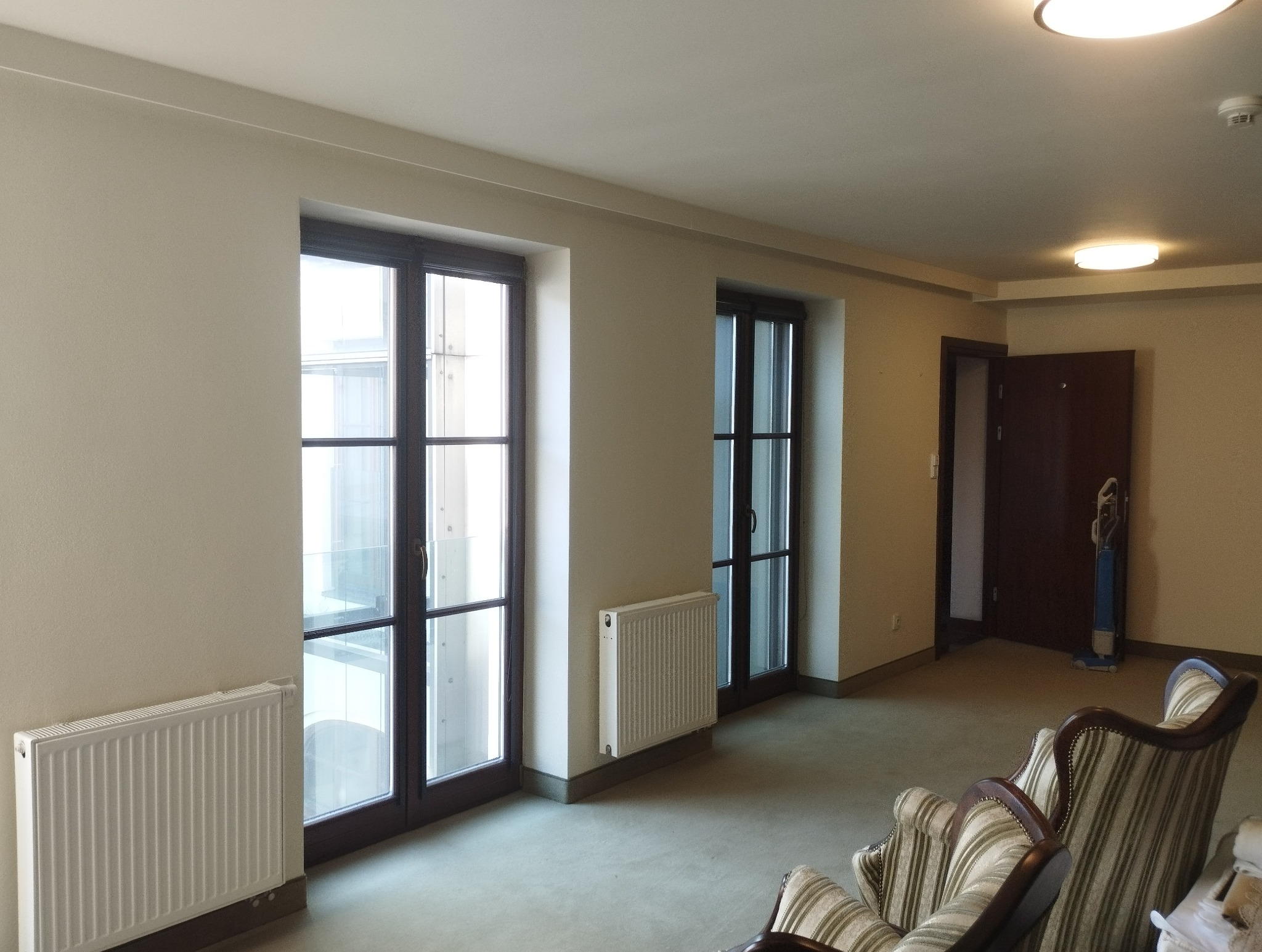 Jasne wnętrze po odświeżeniu: kremowe ściany, ciemne ramy okien, białe grzejniki. Widoczny odkurzacz i fragment fotela w tle. Neutralne oświetlenie.