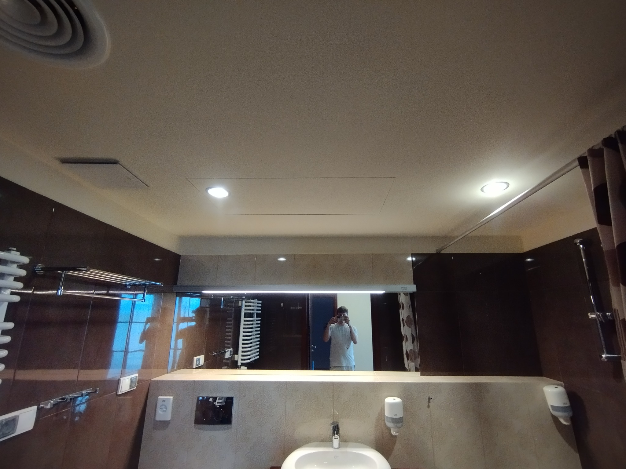 Wykończona łazienka z brązowymi płytkami, białą umywalką i dużym lustrem, w którym odbija się osoba robiąca zdjęcie. Widoczne oświetlenie sufitowe i akcesoria łazienkowe.